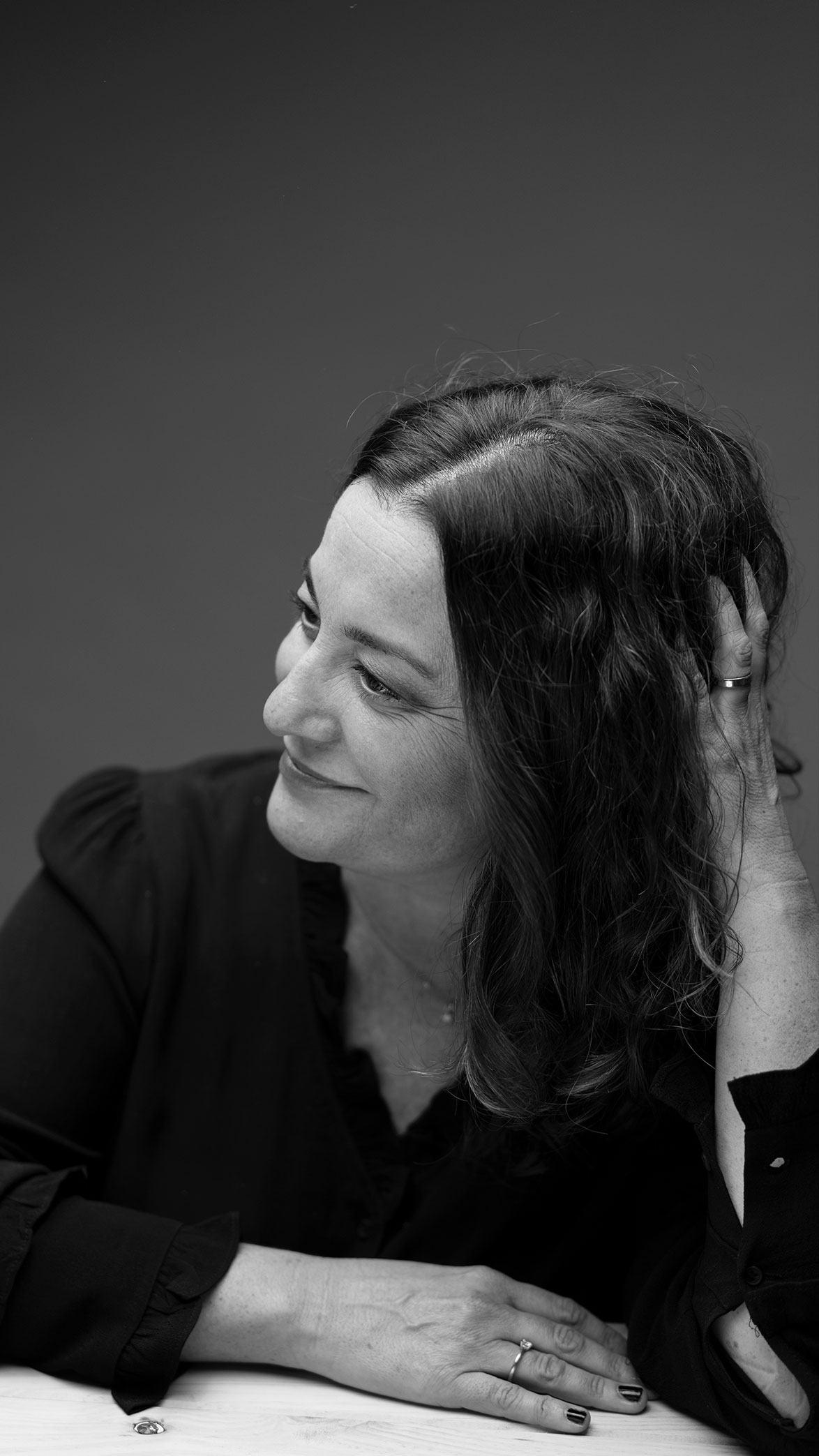 Retrato en blanco y negro de una mujer sonriente con cabello rizado apoyada en una mesa, mirando hacia la izquierda y tocándose el cabello.