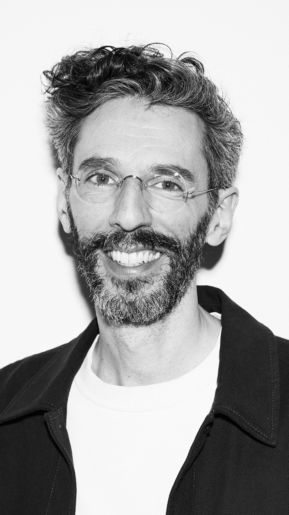 Hombre con gafas redondas, barba canosa y cabello rizado sonriendo, con camiseta blanca y chaqueta oscura.