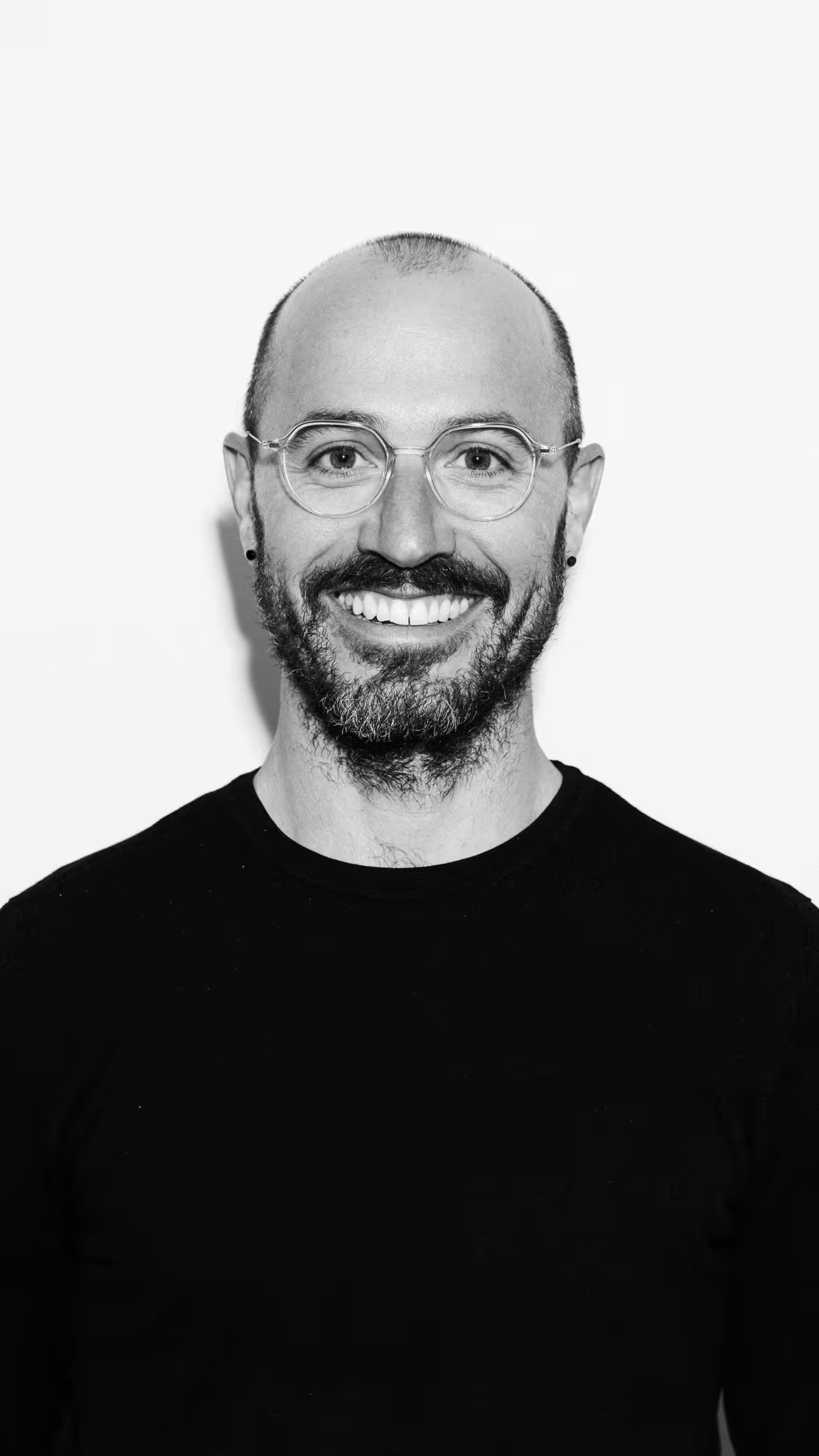Hombre calvo con barba y gafas transparentes sonriendo con camiseta negra frente a fondo blanco.
