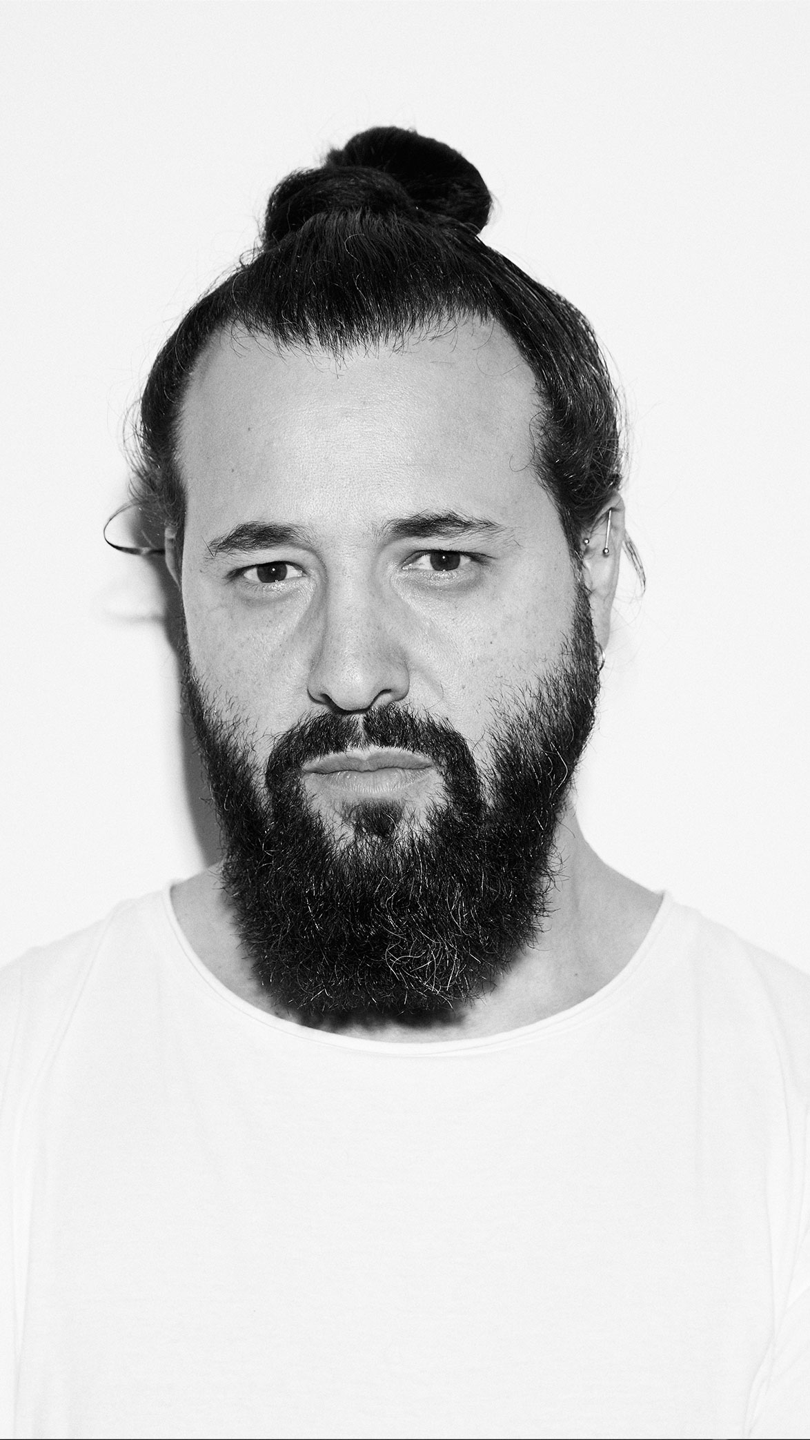 Retrato en blanco y negro de un hombre con barba espesa y cabello recogido en un moño, vistiendo una camiseta blanca.