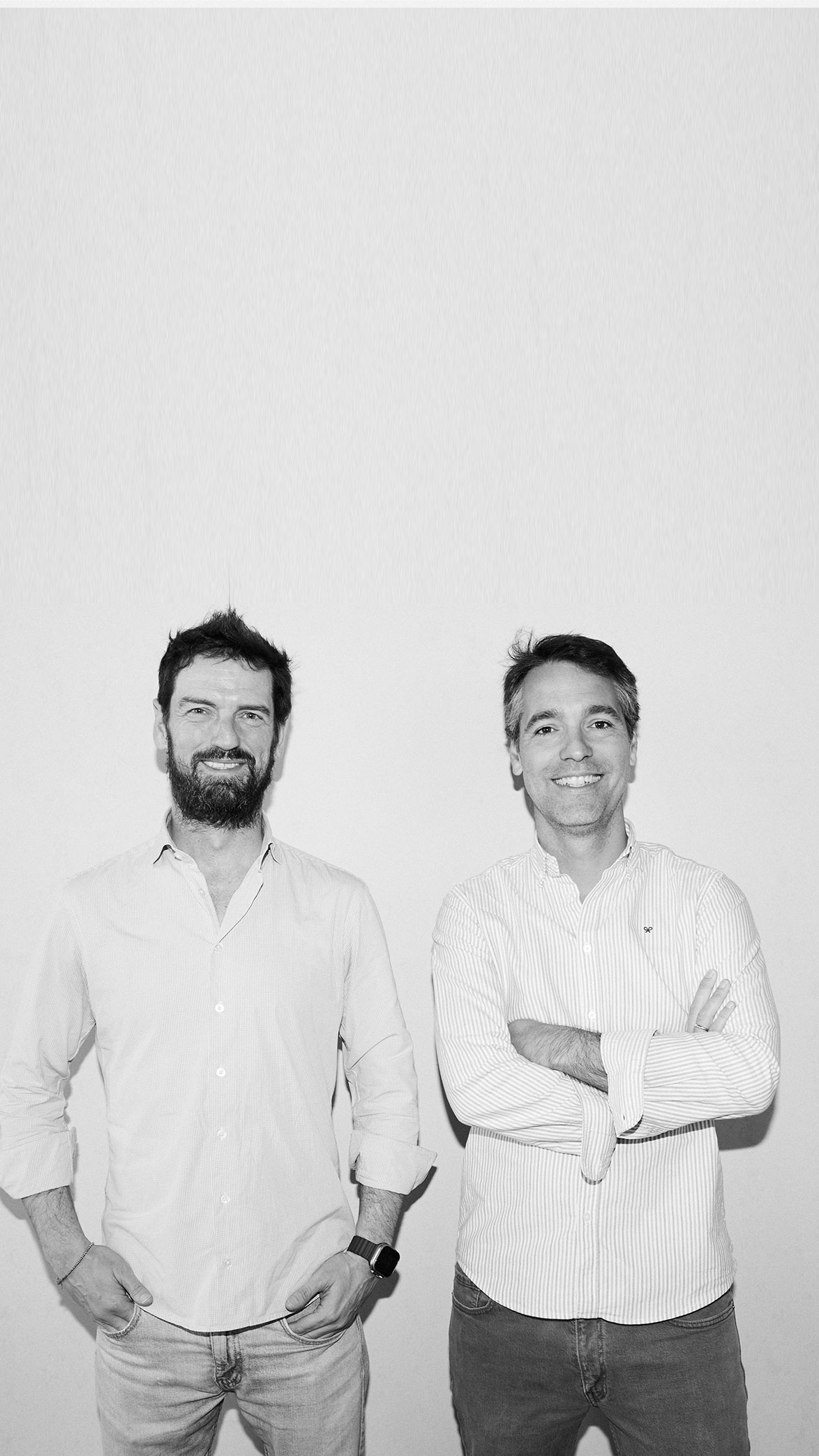 Dos hombres sonrientes de pie frente a una pared blanca, uno con barba y la camisa remangada, el otro con brazos cruzados y camisa de rayas.