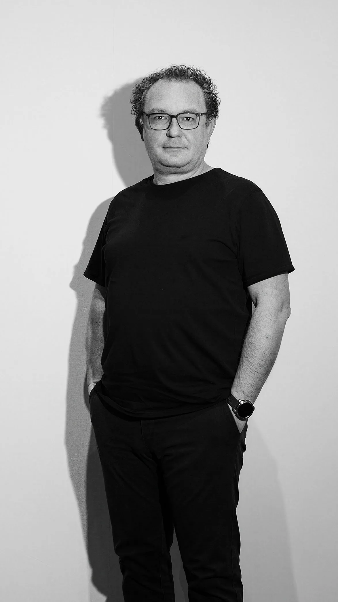 Hombre de mediana edad con gafas, camiseta negra y reloj posando de pie contra una pared clara.
