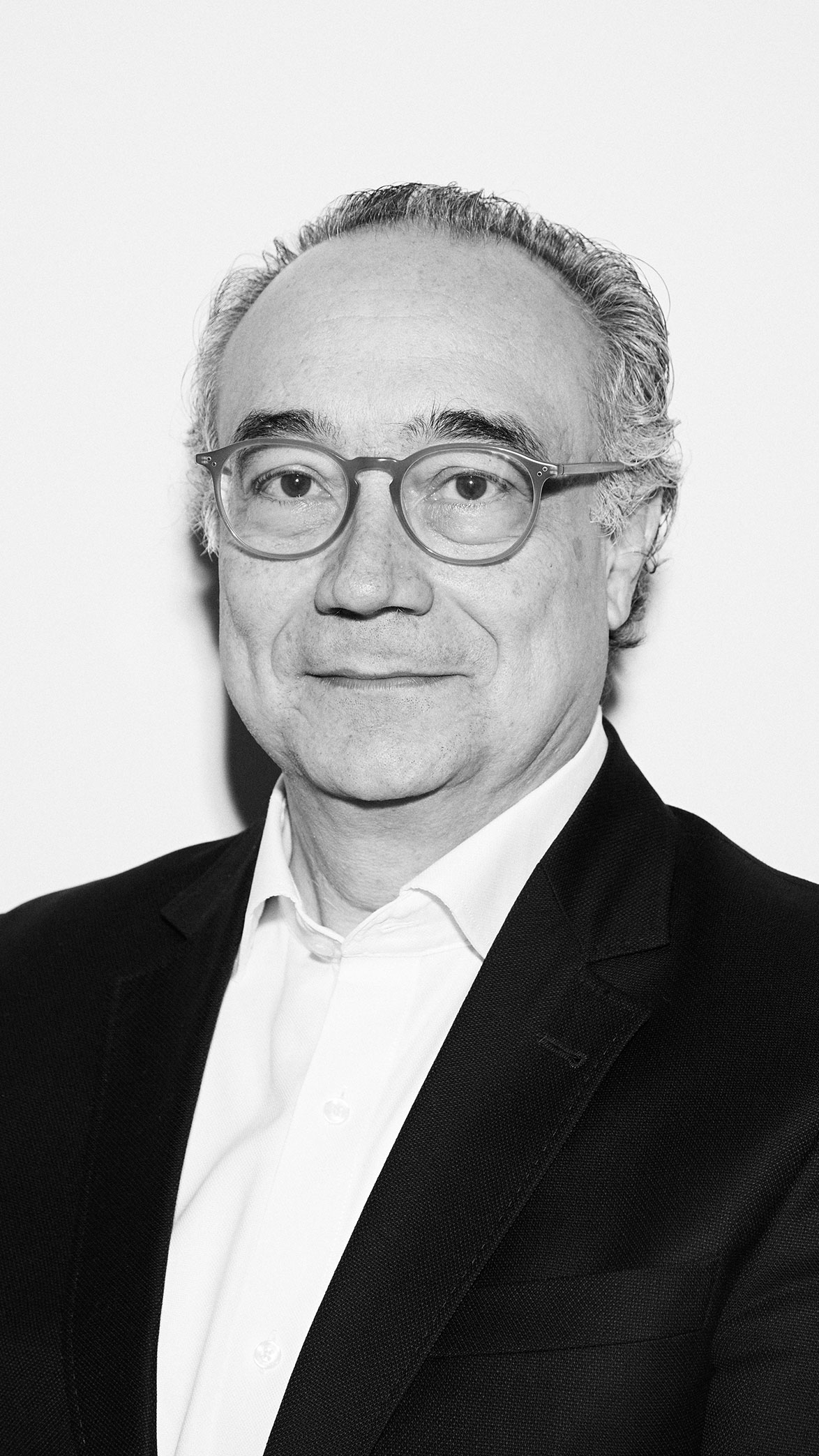 Hombre de mediana edad con gafas y cabello canoso, vestido con camisa blanca y chaqueta negra, posando frente a fondo blanco.