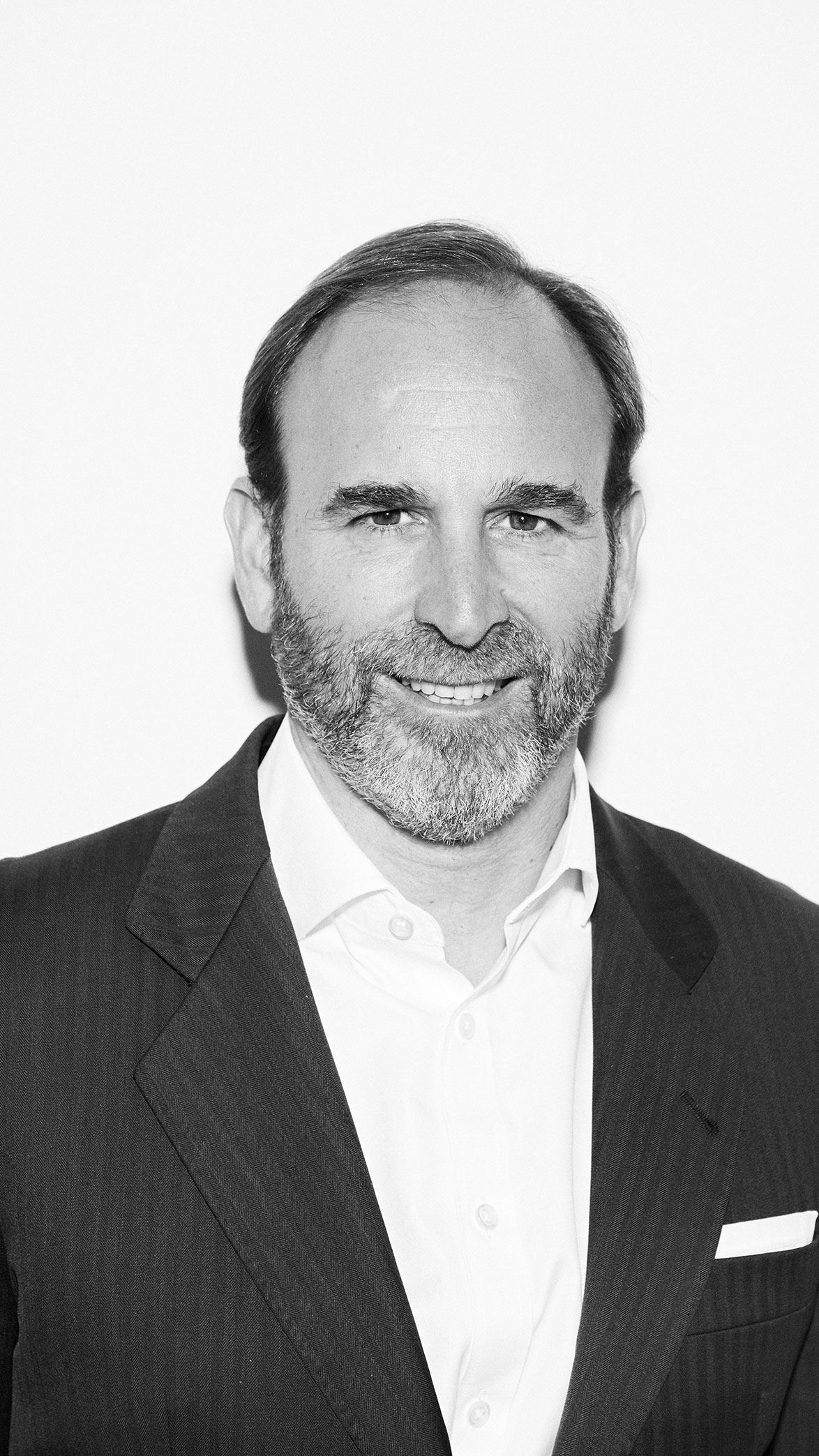 Hombre con barba y bigote sonriendo, vestido con traje oscuro y camisa blanca.