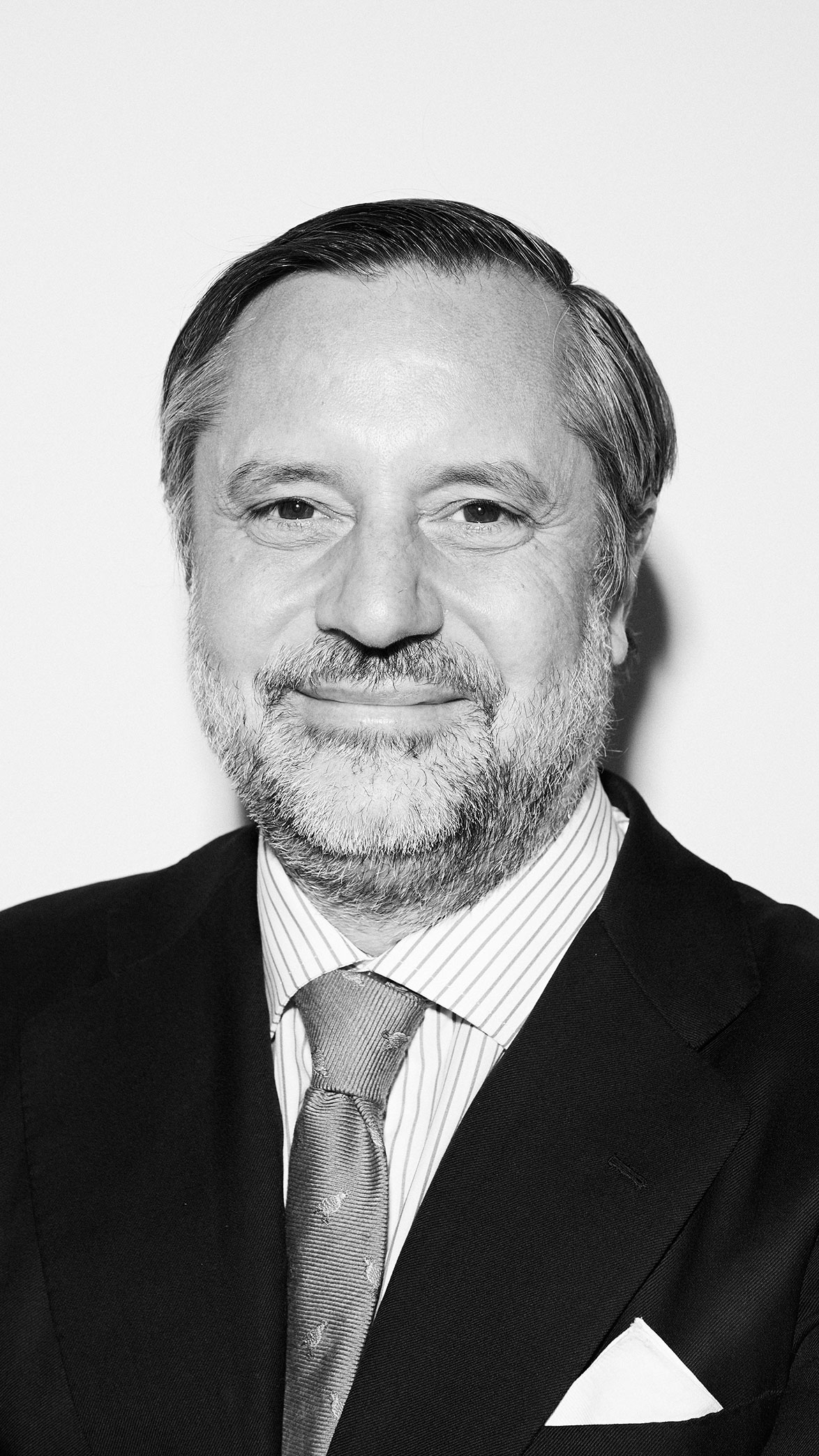 Hombre de mediana edad con barba y cabello corto, vestido con traje oscuro, camisa a rayas y corbata, sonriendo levemente.