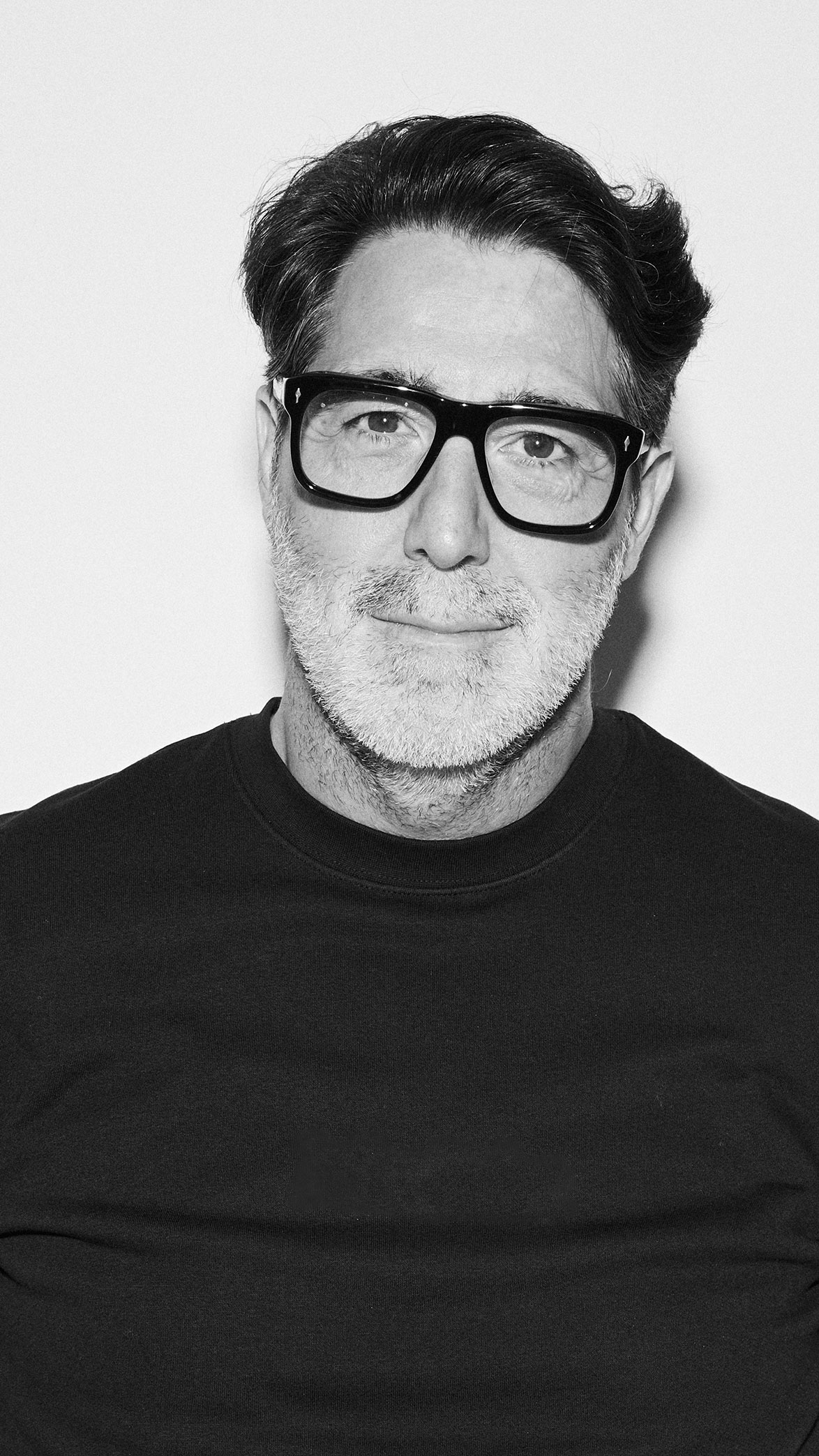 Hombre con gafas negras grandes, barba canosa y camiseta negra sonriendo levemente en fondo blanco.