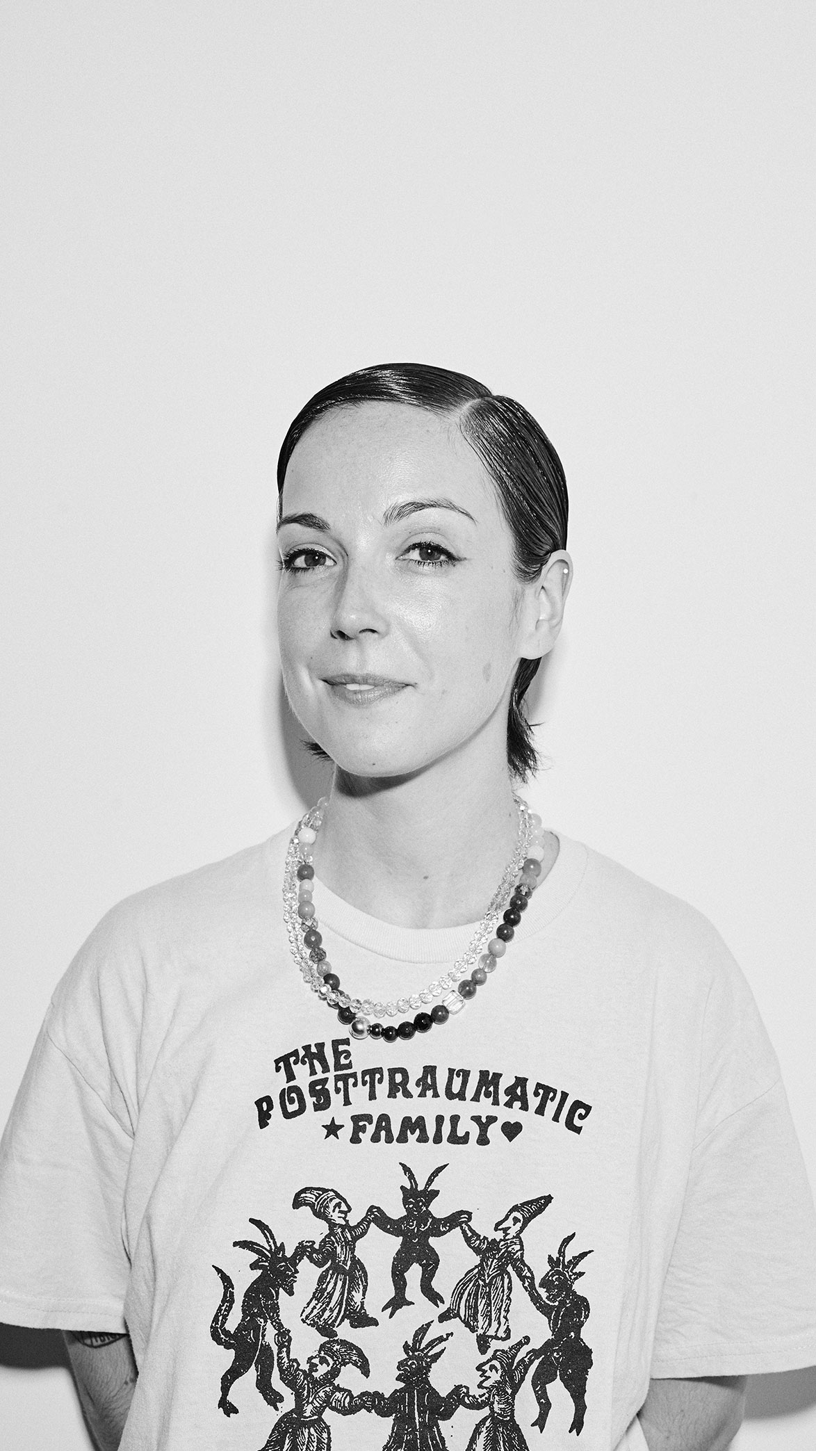 Retrato en blanco y negro de una mujer con cabello corto peinado hacia atrás, usando una camiseta con el texto 'The Posttraumatic Family' y un collar de cuentas.