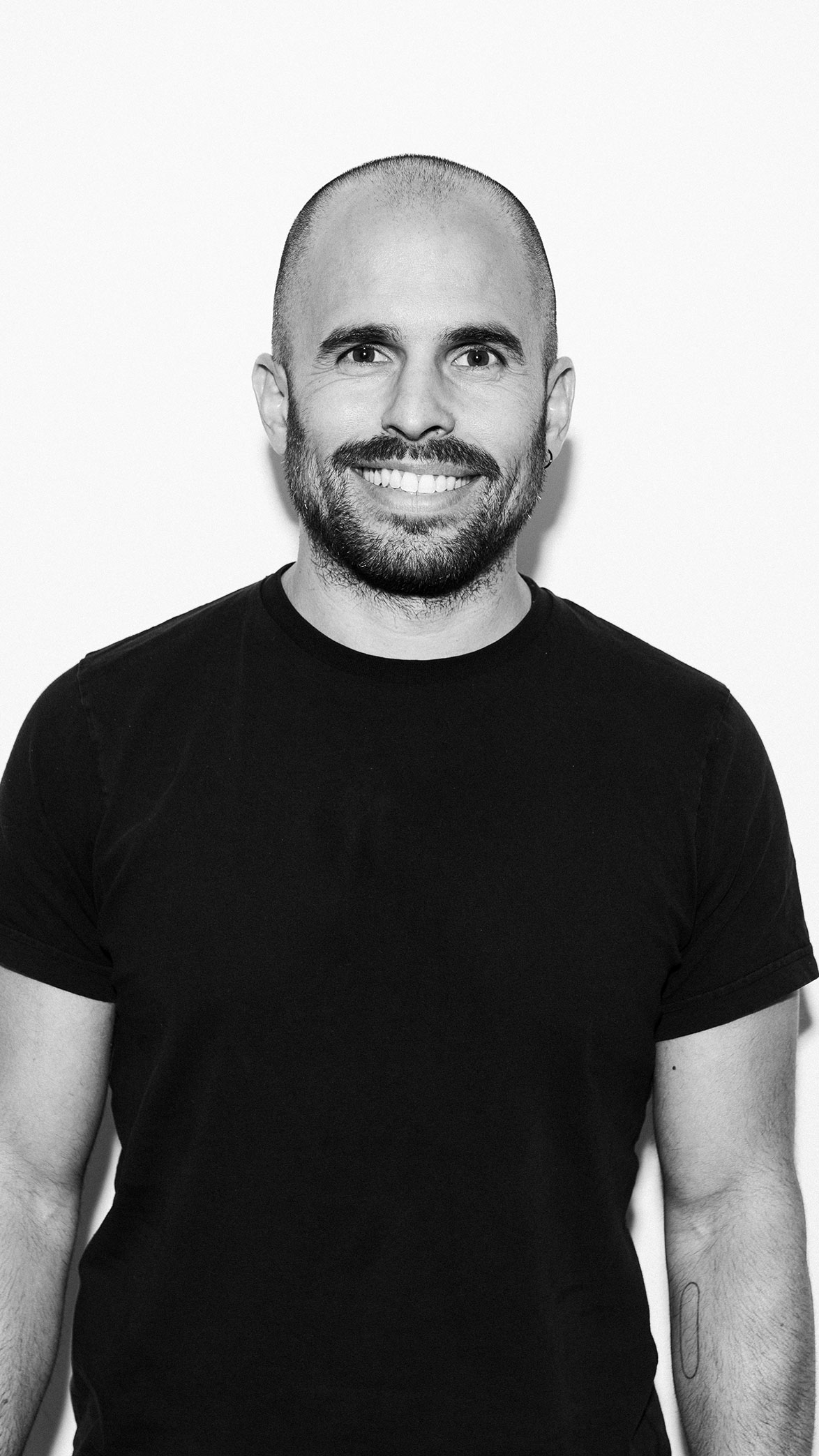 Hombre calvo con barba y camiseta negra sonriente frente a fondo blanco.