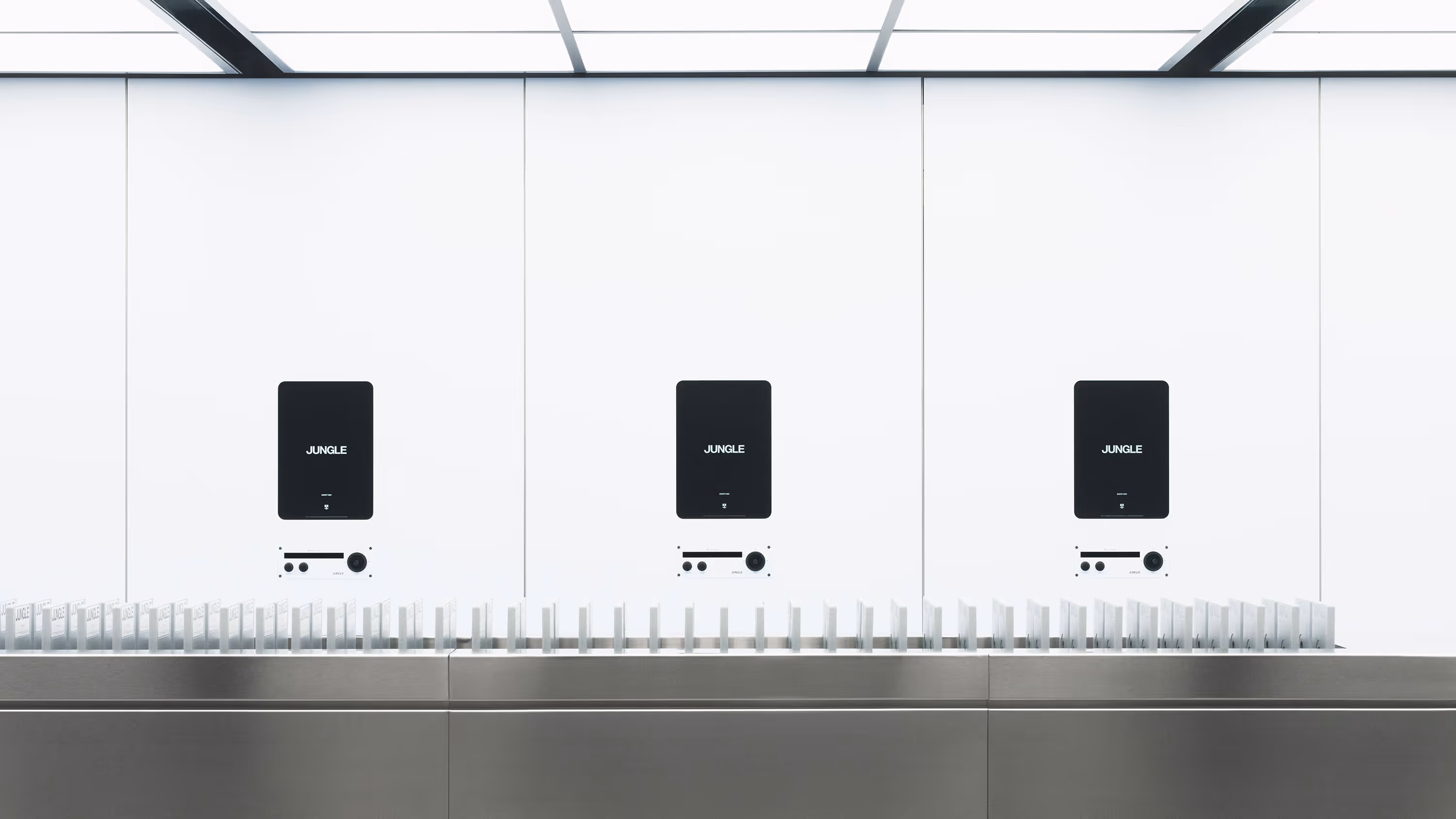 Espacio minimalista con tres pantallas negras sobre fondo blanco, cada una con la palabra 'JUNGLE', y una fila de objetos blancos alineados en una superficie metálica.