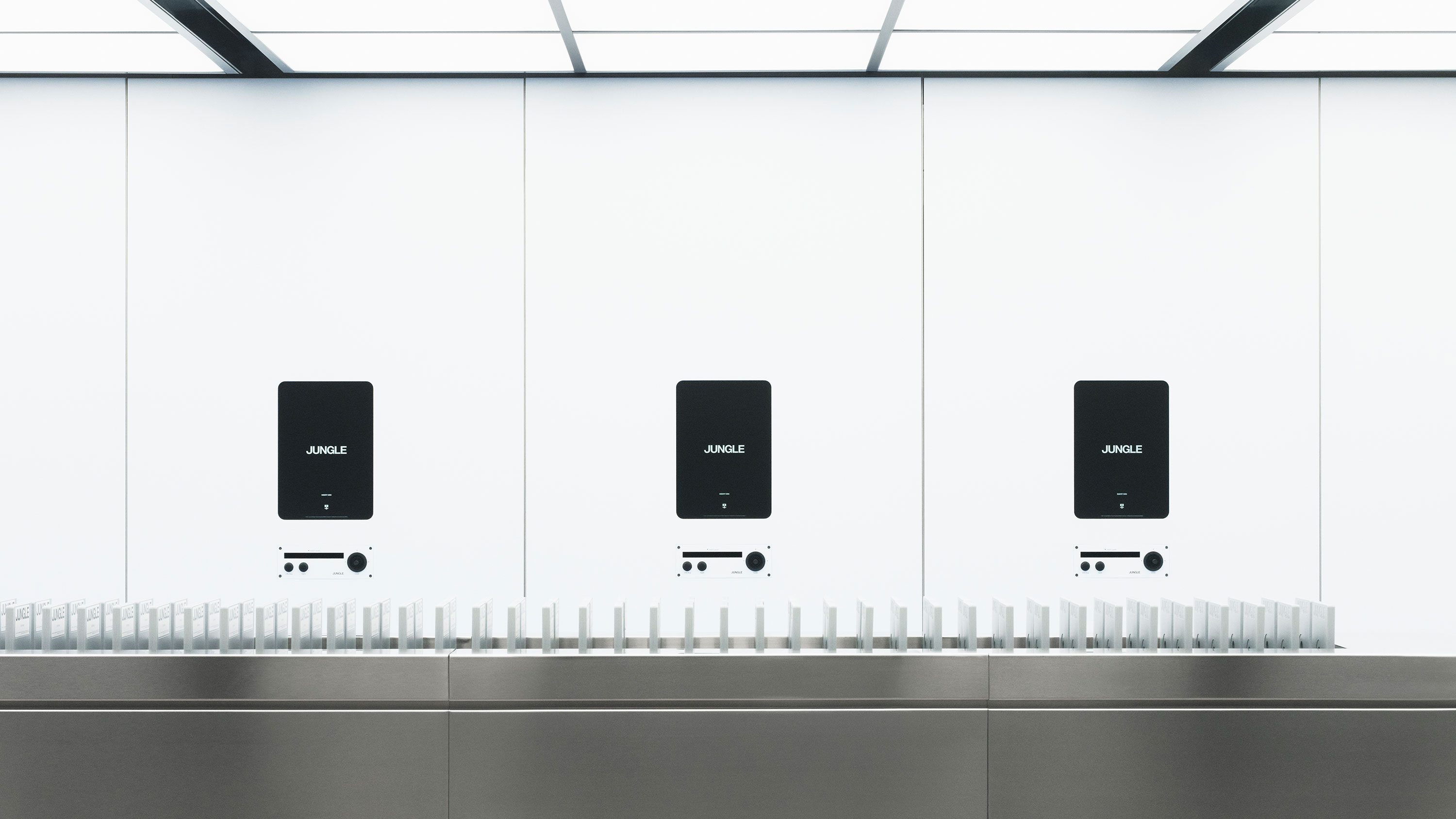 Espacio minimalista con tres pantallas negras sobre fondo blanco, cada una con la palabra 'JUNGLE', y una fila de objetos blancos alineados en una superficie metálica.