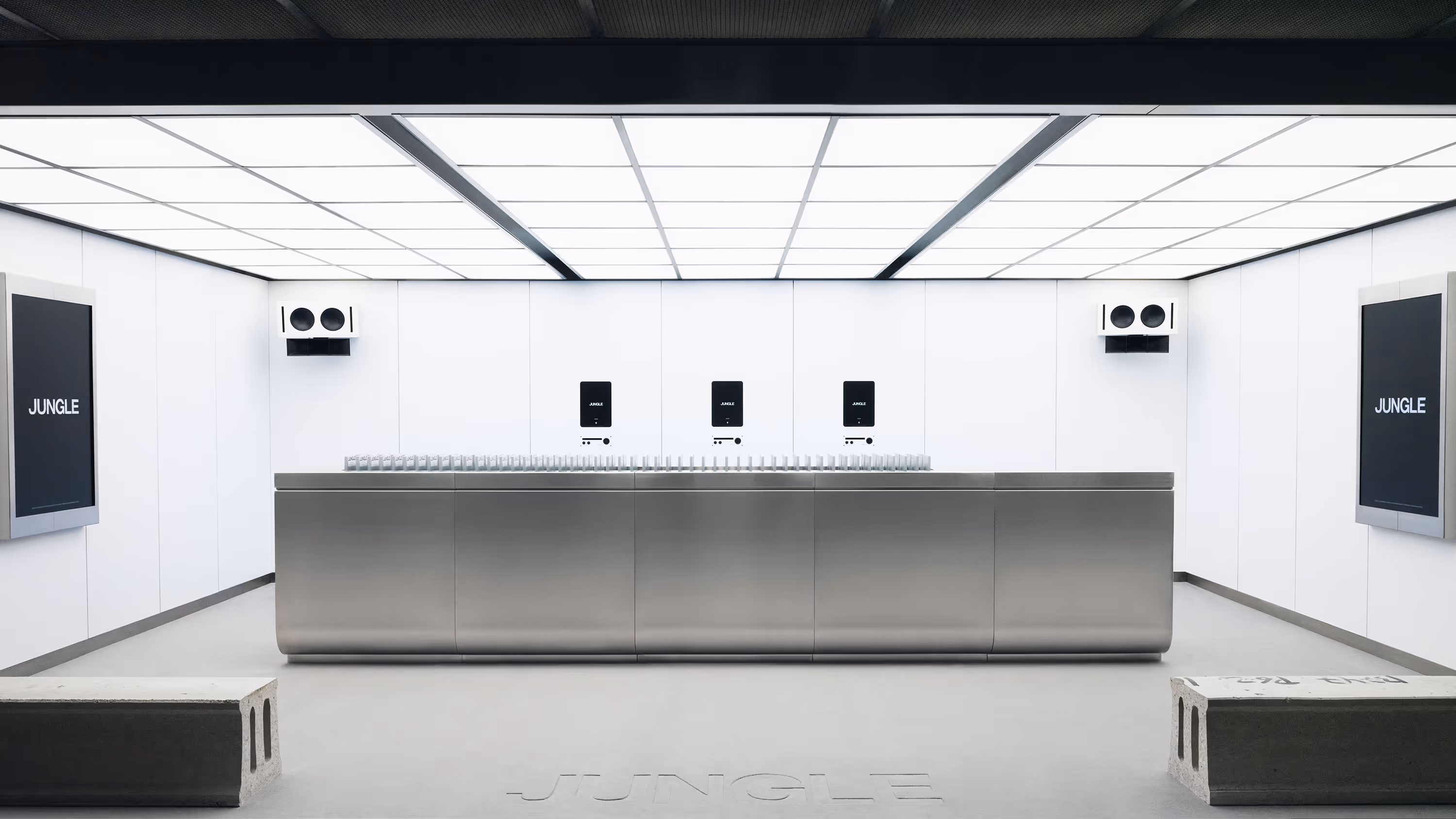 Mostrador metálico en una sala blanca minimalista con letreros que dicen 'JUNGLE' y paneles de iluminación en el techo.