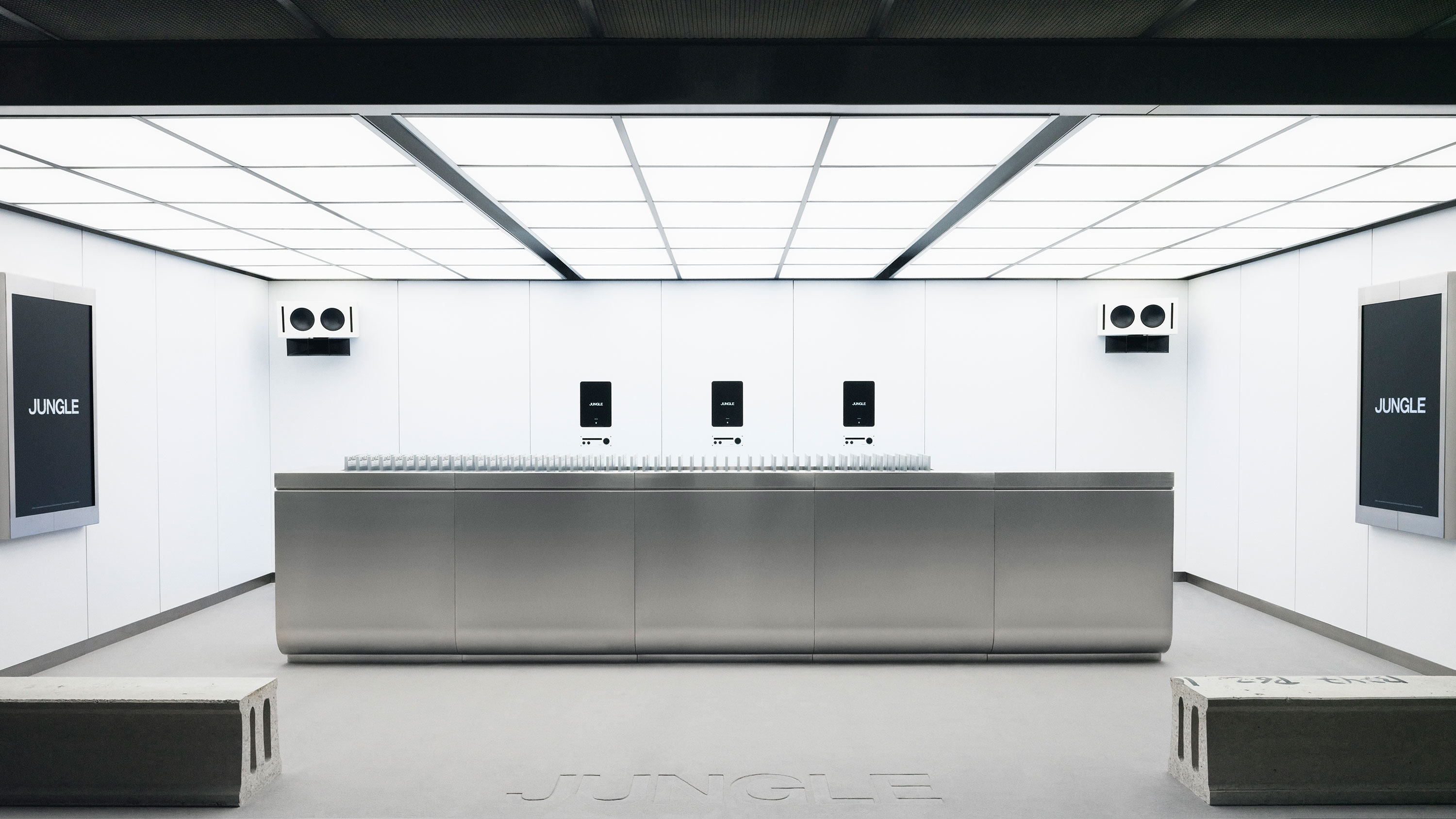 Mostrador metálico en una sala blanca minimalista con letreros que dicen 'JUNGLE' y paneles de iluminación en el techo.