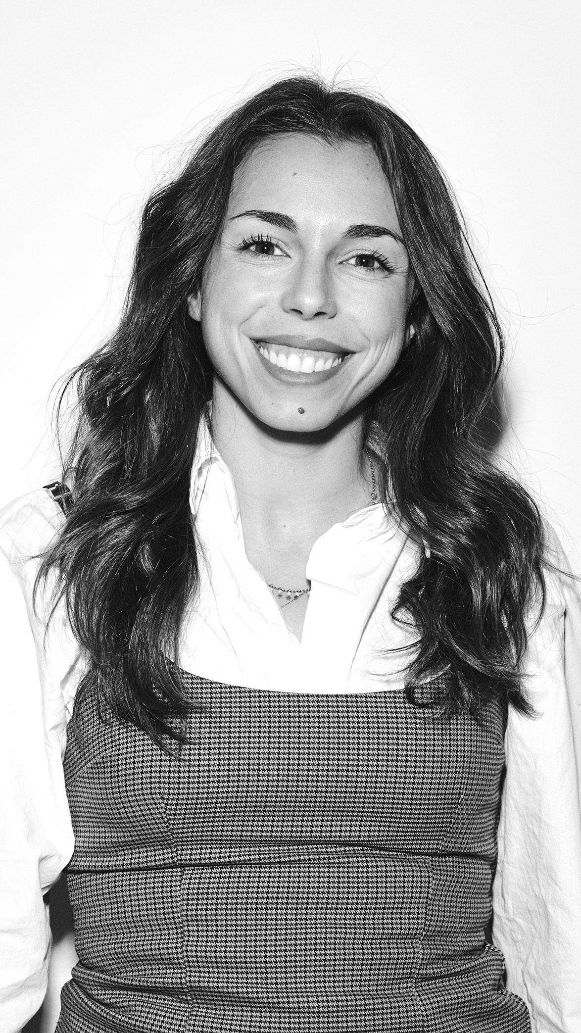 Retrato en blanco y negro de una mujer sonriente con cabello ondulado, camisa blanca y vestido de patrón de cuadros.