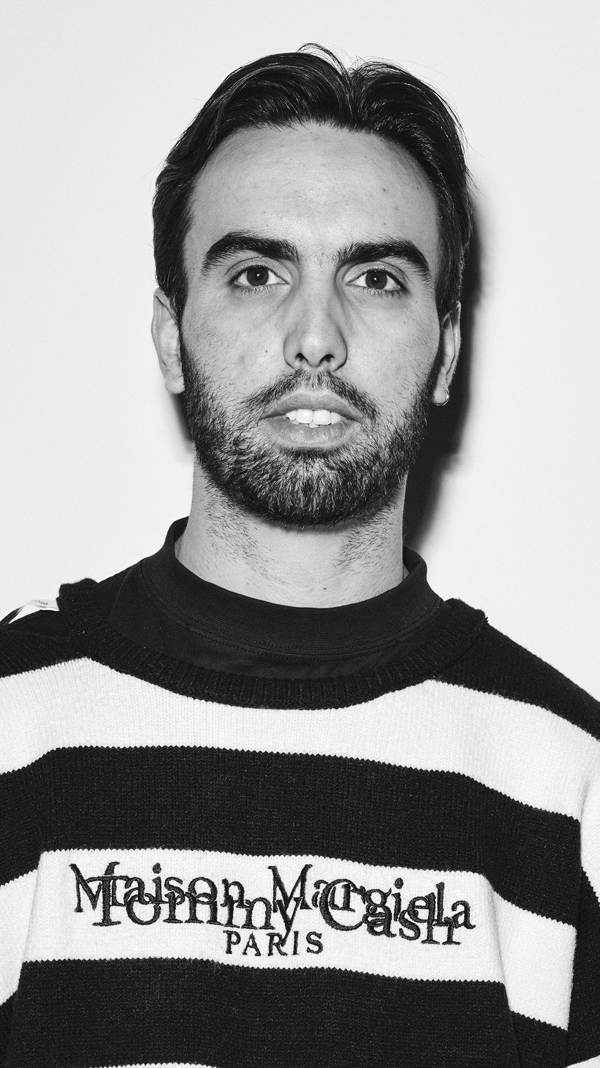 Retrato en blanco y negro de un hombre con barba y cabello oscuro corto, vistiendo un suéter de rayas blancas y negras con el texto Maison Margiela Paris.