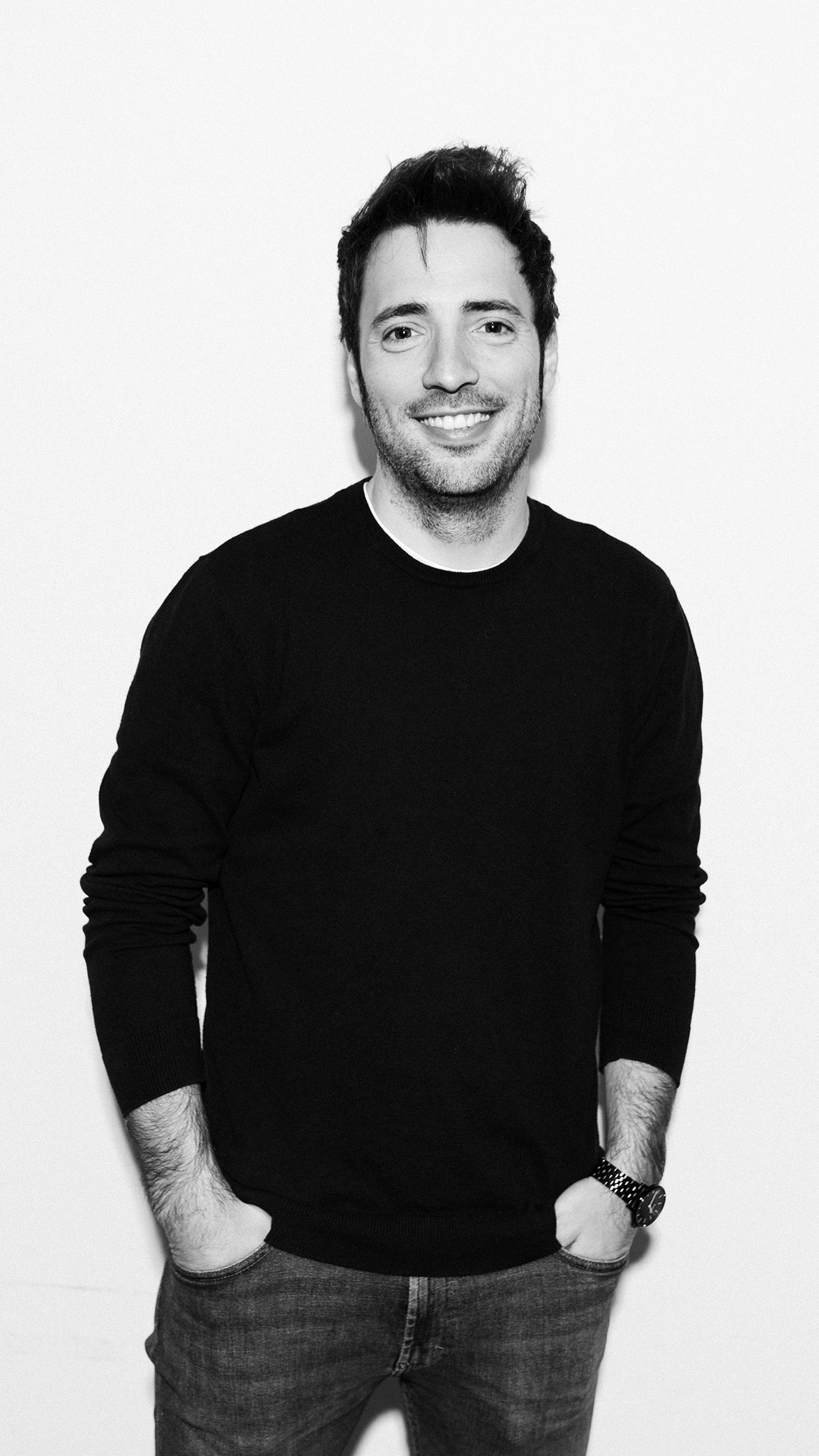Hombre joven sonriente con suéter negro y jeans, con las manos en los bolsillos, posando frente a fondo blanco.