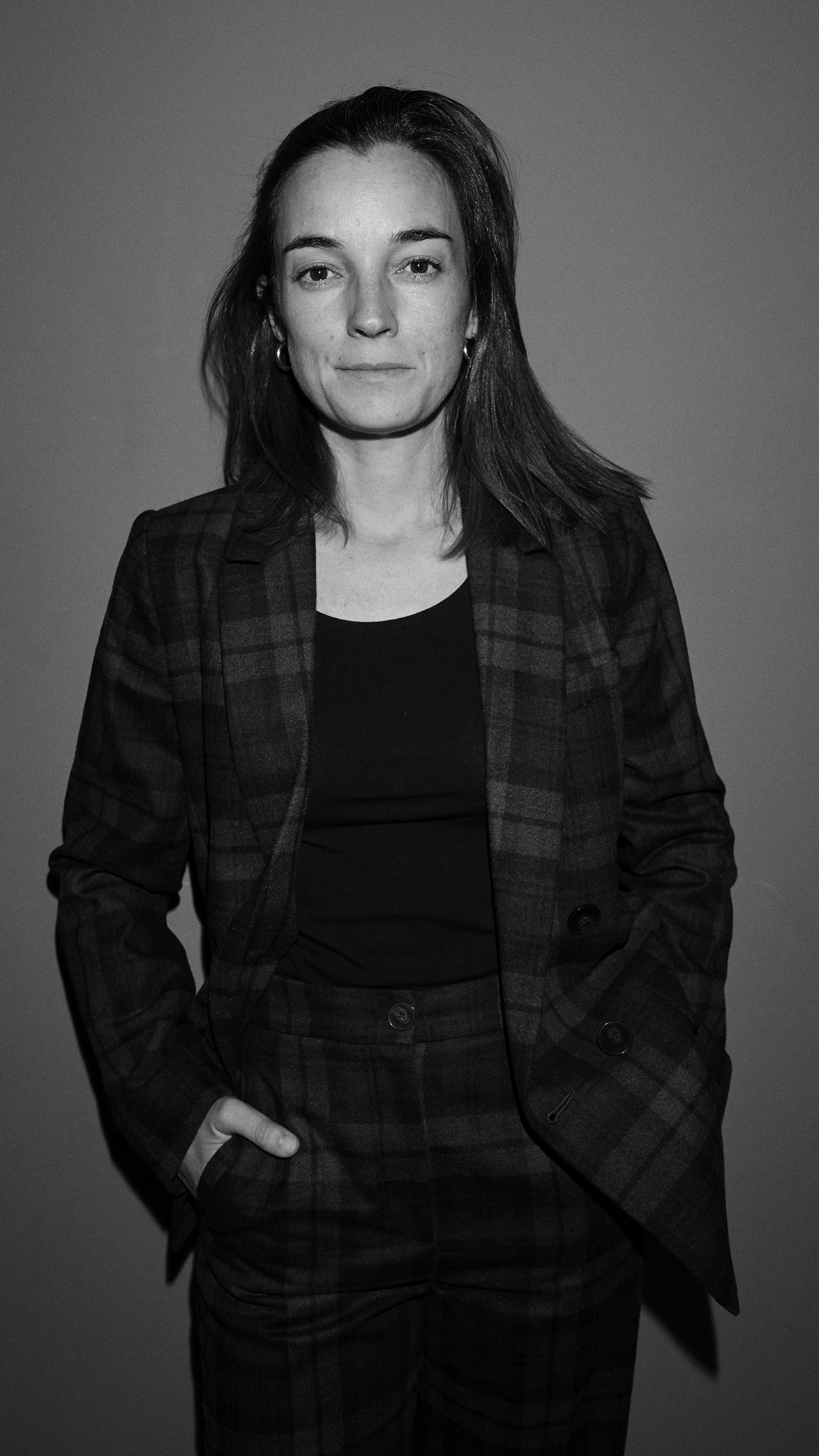 Mujer con chaqueta y pantalones de cuadros oscuros y camiseta negra, con manos en los bolsillos, mirando al frente en fondo gris.