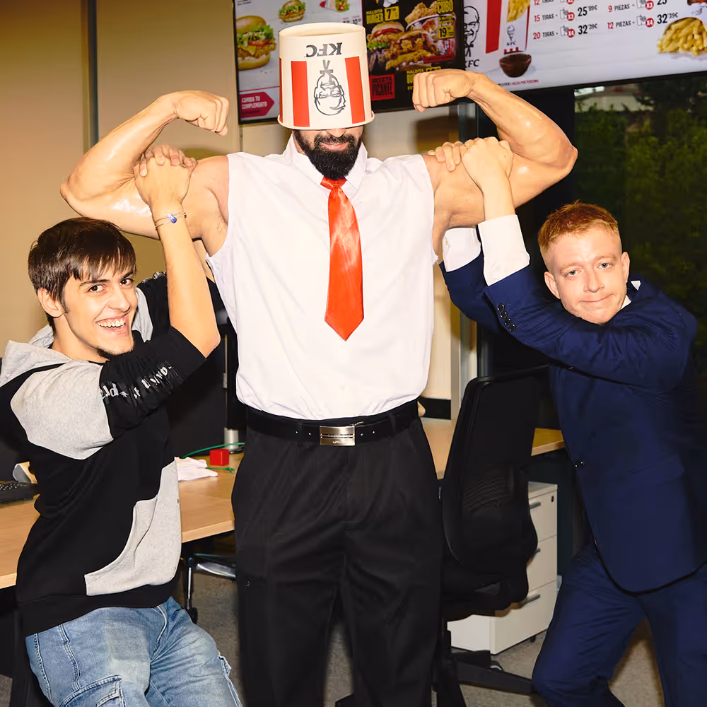 Hombre fuerte con un cubo de KFC en la cabeza y corbata roja flexionando brazos mientras dos hombres lo sujetan y sonríen en una oficina.