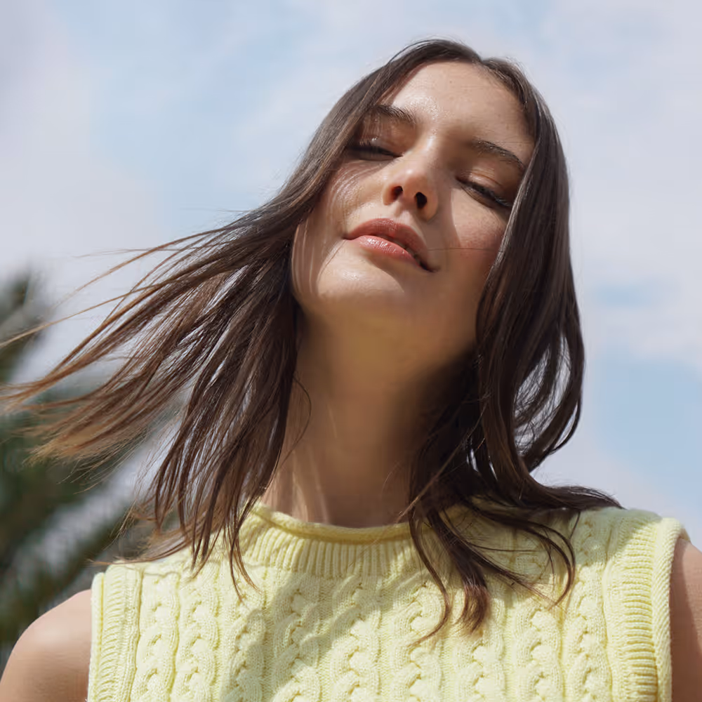 Primer plano de una mujer con cabello castaño y suelto, ojos cerrados y suéter amarillo trenzado bajo un cielo azul claro.