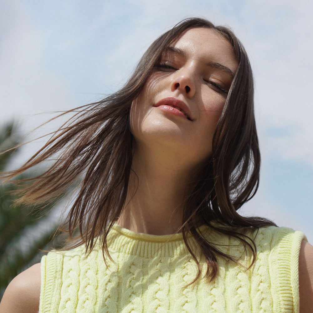 Primer plano de una mujer con cabello castaño y suelto, ojos cerrados y suéter amarillo trenzado bajo un cielo azul claro.
