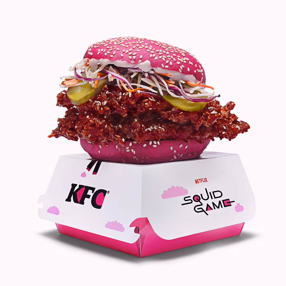 Sándwich de pollo frito con pan rosa y semillas de sésamo, repollo rallado, pepinillos y salsa, sobre caja de KFC temática de Squid Game.