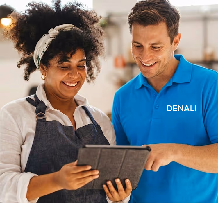 Smiling woman in apron and man in blue shirt looking at a tablet together indoors.