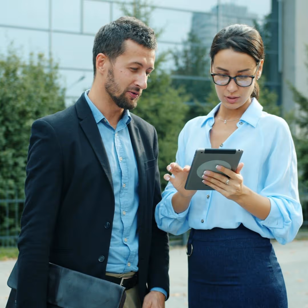 Man and woman dressed in business attire looking at a tablet outdoors near a modern building.