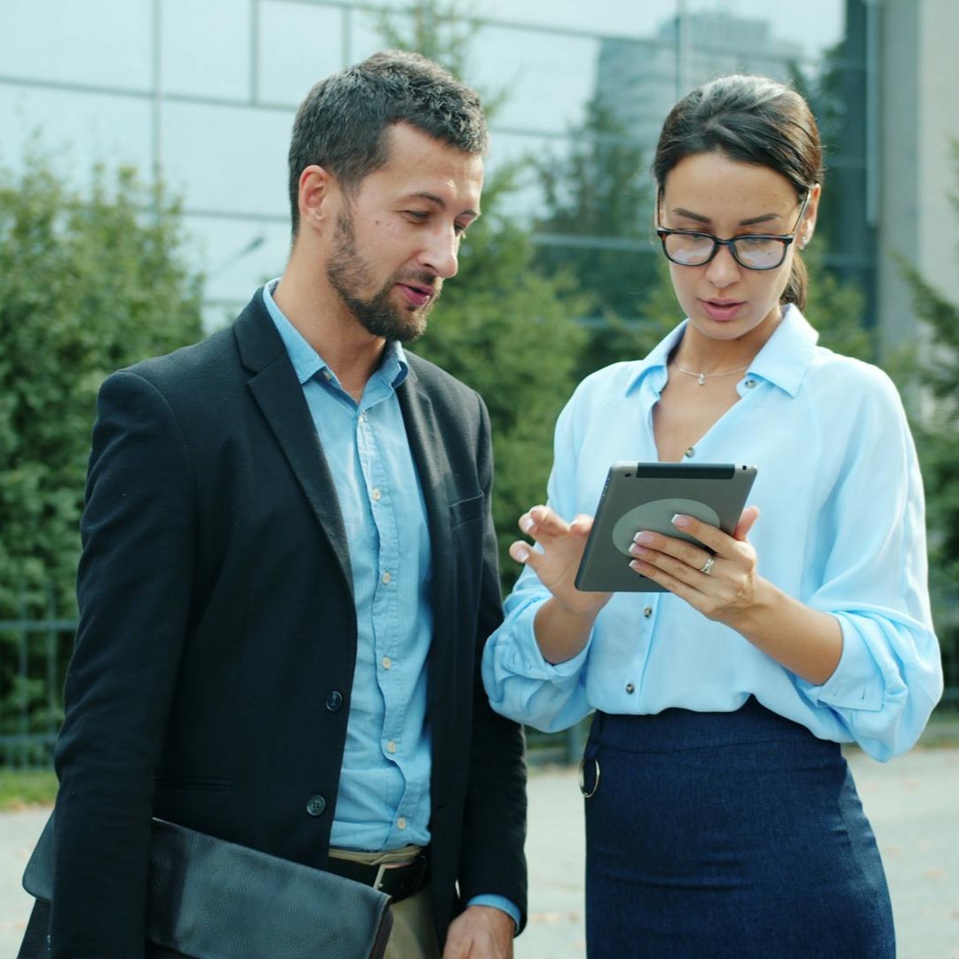 Man and woman dressed in business attire looking at a tablet outdoors near a modern building.