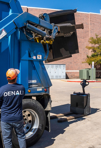 Worker in Denali shirt operates machinery lifting a black container outdoors near a brick building.