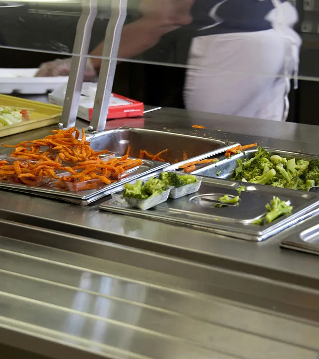 Cafeteria food tray with sweet potato fries and steamed broccoli in metal serving containers.