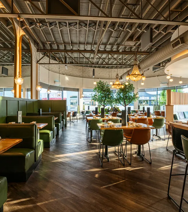 Spacious modern restaurant interior with green booths, wooden tables, chandeliers, and decorative indoor trees.