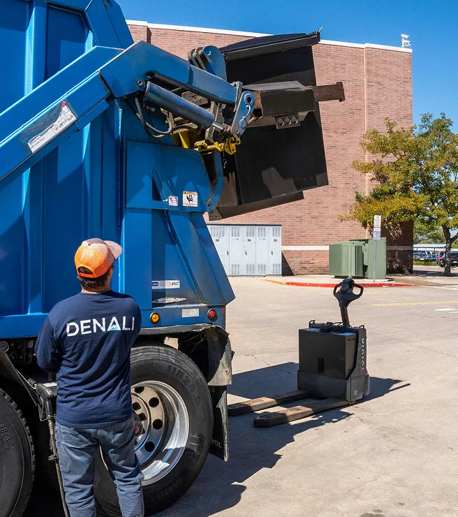 Worker with Denali shirt operating blue garbage truck lifting a black bin outside a brick building.