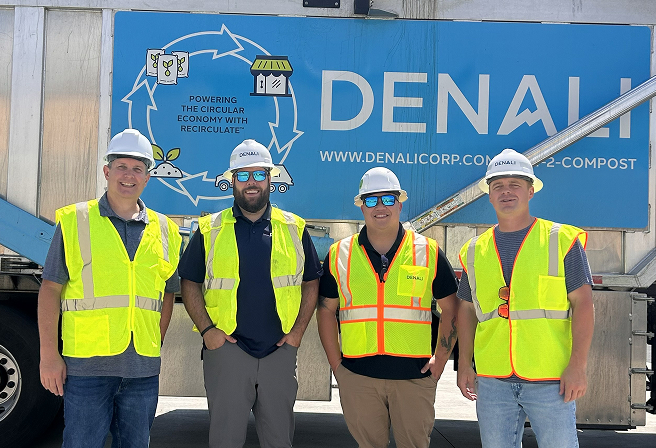 Four men wearing white hard hats and yellow safety vests standing in front of a large truck with a blue Denali banner displaying a circular economy illustration and the text 'Powering the circular economy with recirculate'.