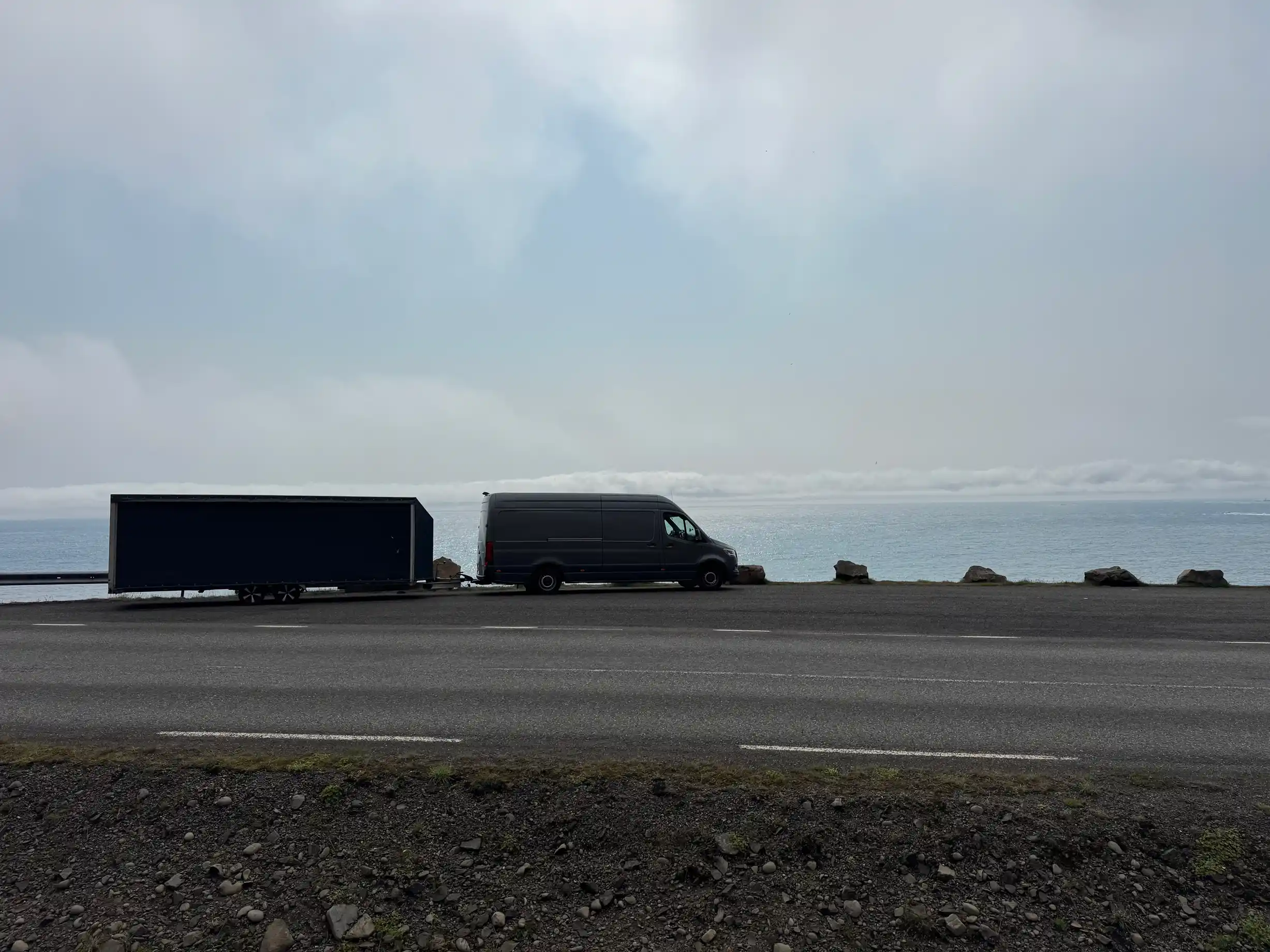Grey van and enclosed trailer parked on the roadside, with the sea in the background.