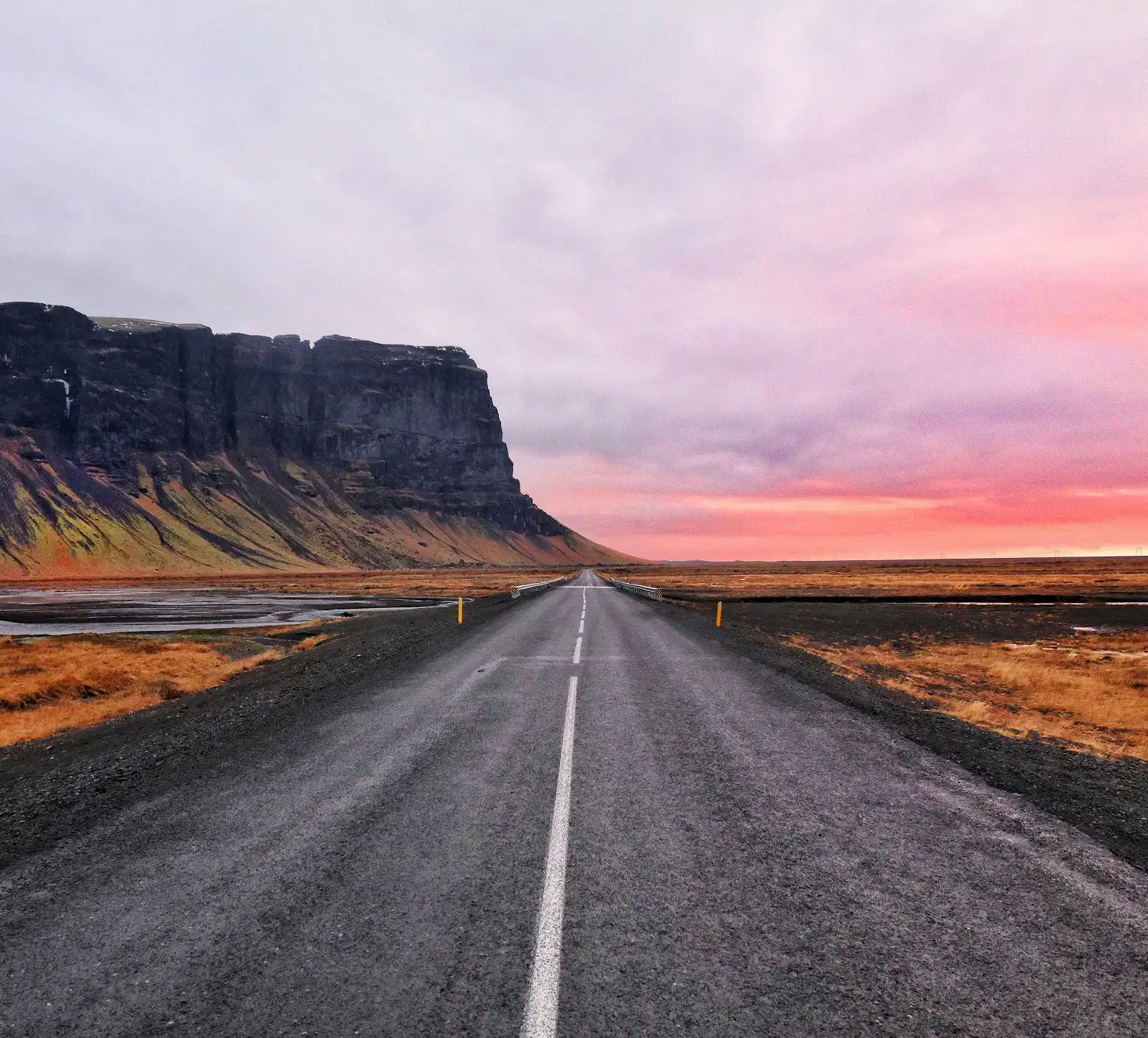 Straight asphalt road is running next to a rocky mountain in the sunset.
