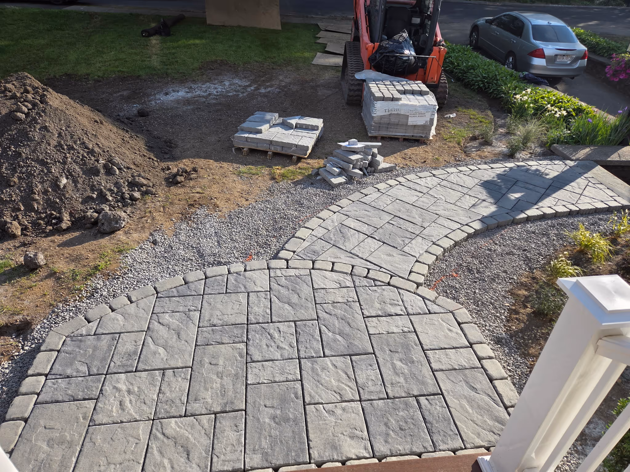 Partially completed stone patio and walkway under construction with stacks of paving stones, gravel, and a small orange skid steer loader nearby.