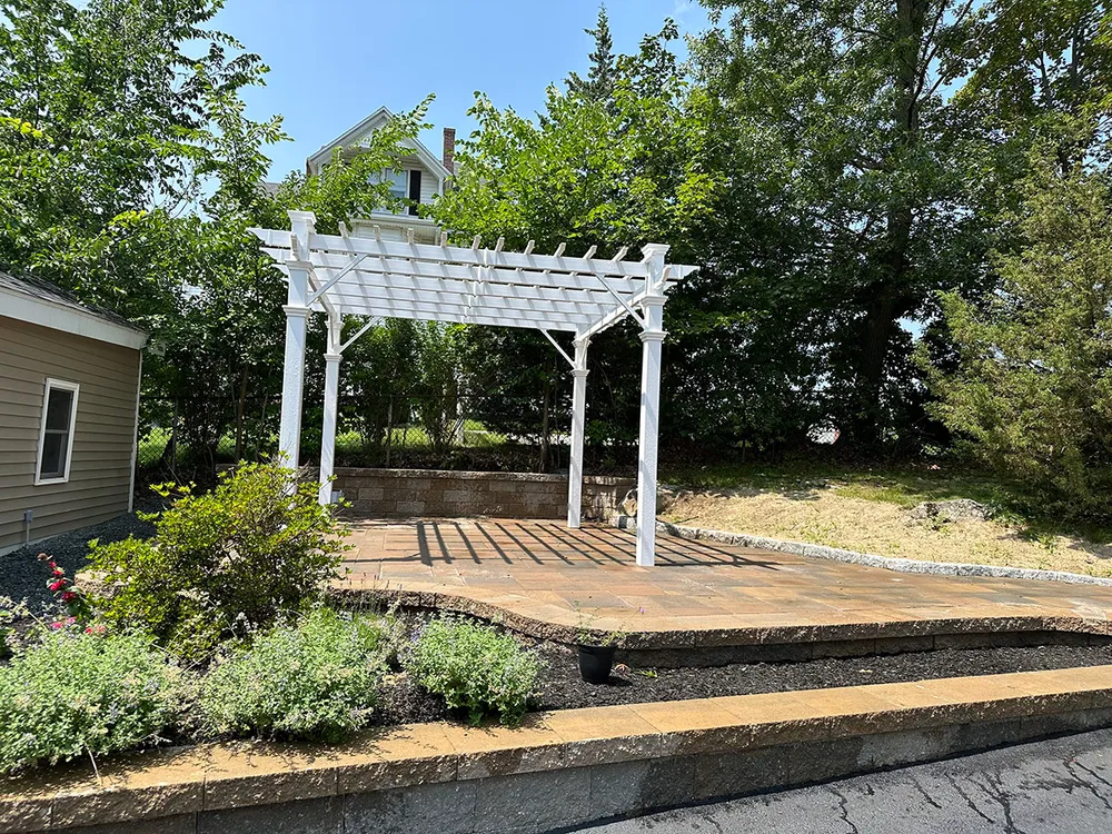 White wooden pergola casting shadows on a stone patio surrounded by greenery and shrubs under a clear blue sky.