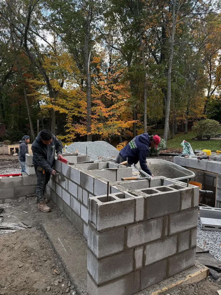 Two construction workers building a cinder block wall outdoors with fall-colored trees in the background.