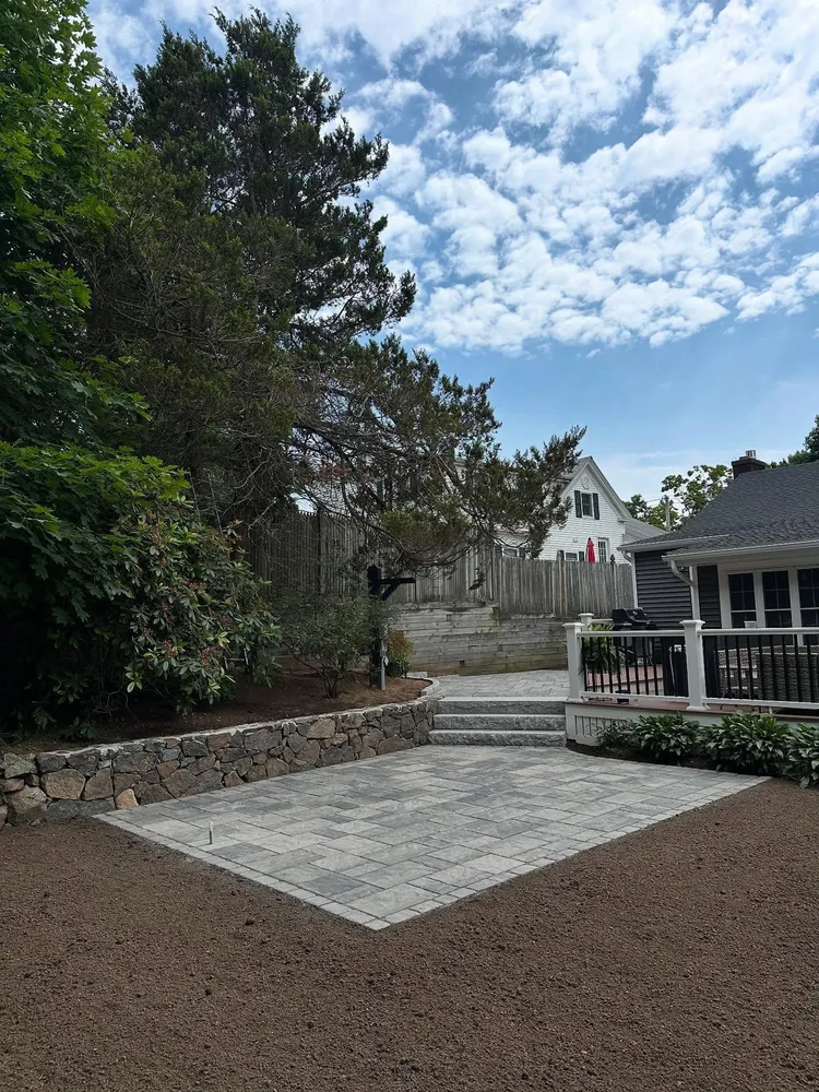 A backyard with a newly paved stone patio, surrounded by garden soil, plants, and trees, adjacent to a house with a railing.