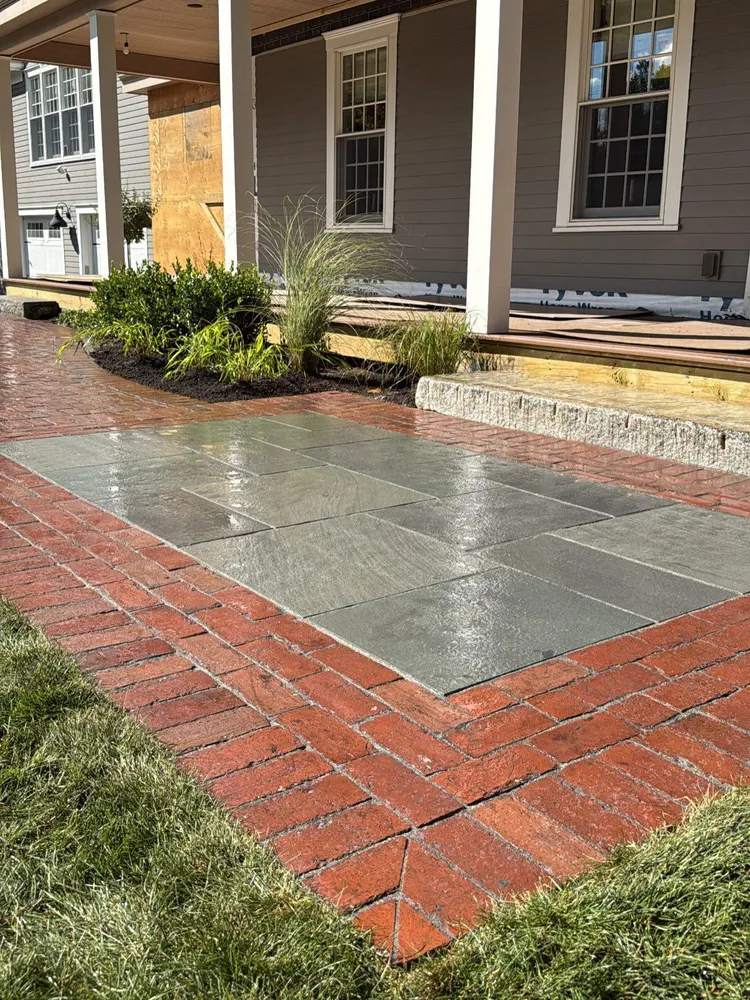 Wet stone patio section bordered by red bricks leading to a porch with green plants and gray siding.