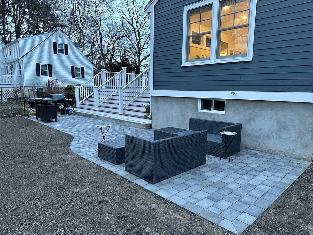 Backyard patio with gray stone pavers, outside wicker furniture, small tables, and stairs leading to a blue house with lit windows.