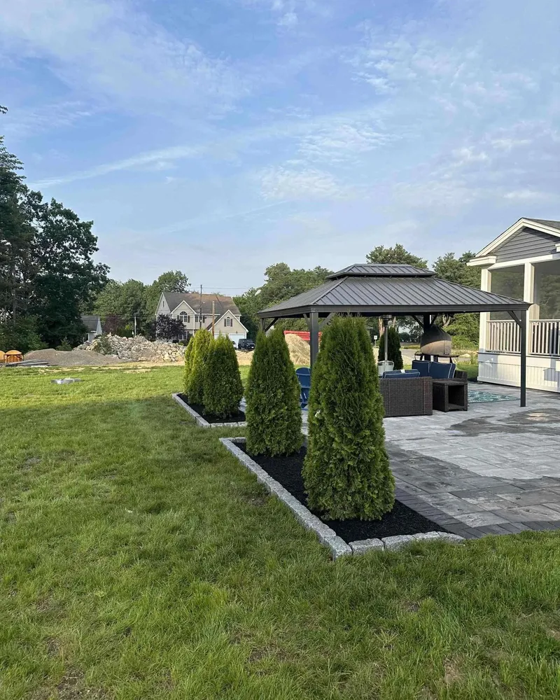 Backyard patio area with a gazebo, outdoor furniture, and a row of small evergreen trees bordered by stone edging and black mulch.