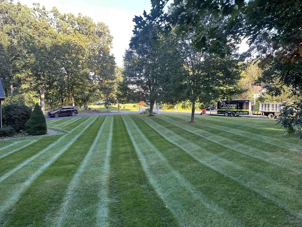 Well-manicured lawn with alternating light and dark green mowing stripes, surrounded by mature trees and a landscaping truck in the background.