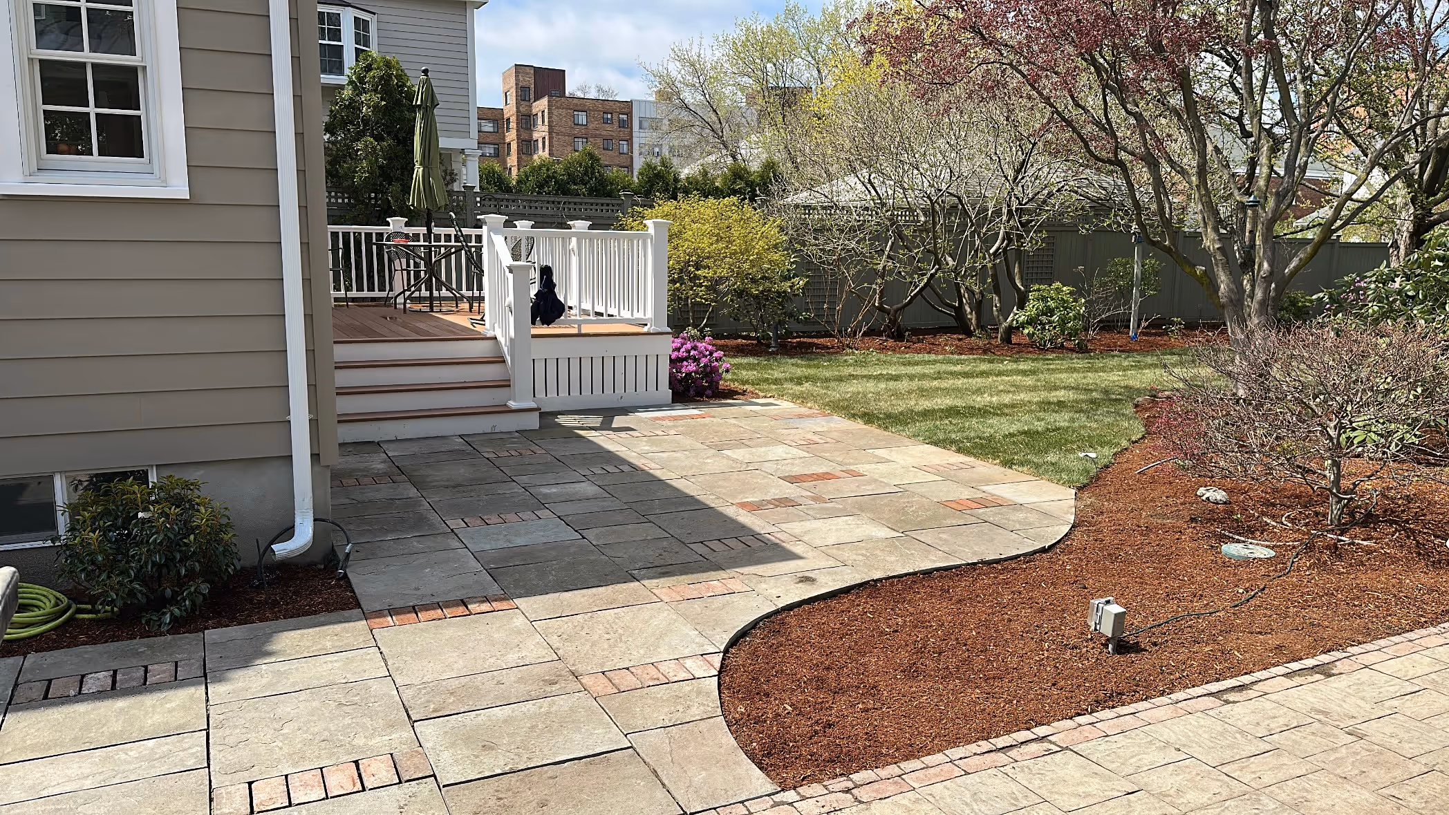 Backyard with a stone patio, small wooden deck with white railing, green lawn, and mulched garden areas with trees and shrubs.