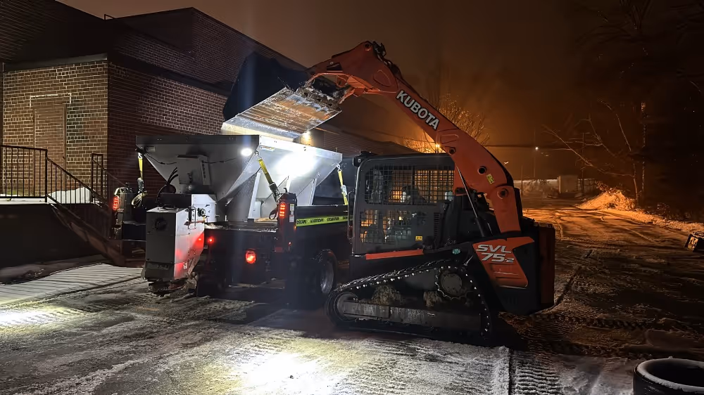 Kubota mini excavator loading material into a truck at night on a snowy path near a brick building.