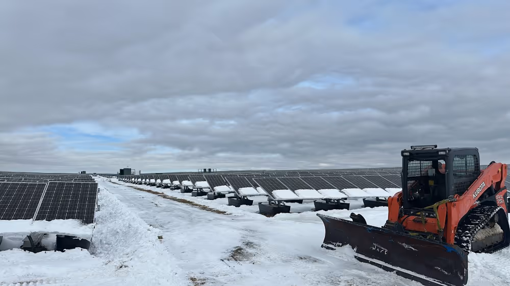 Snow-covered solar panels arranged in rows with a snowplow clearing a path.