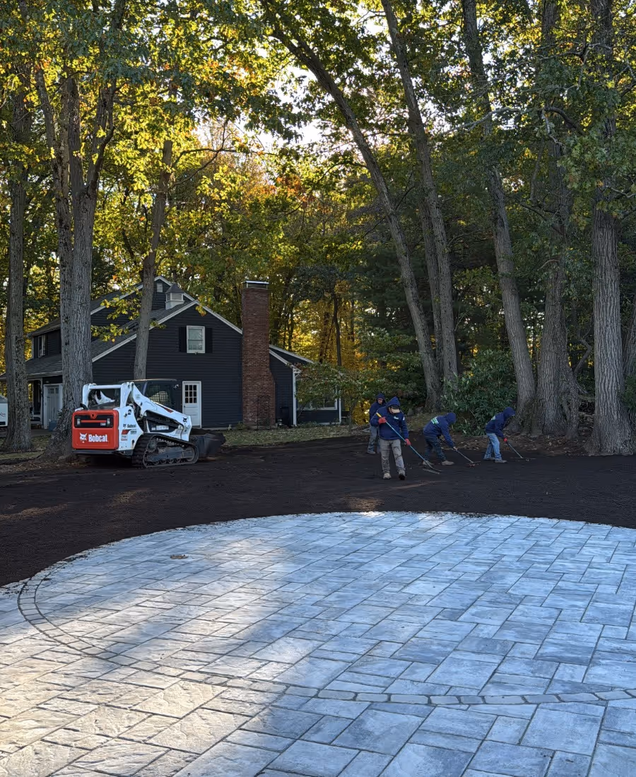 Workers raking soil next to a circular stone patio in a wooded backyard with a dark gray house and a Bobcat skid-steer loader.