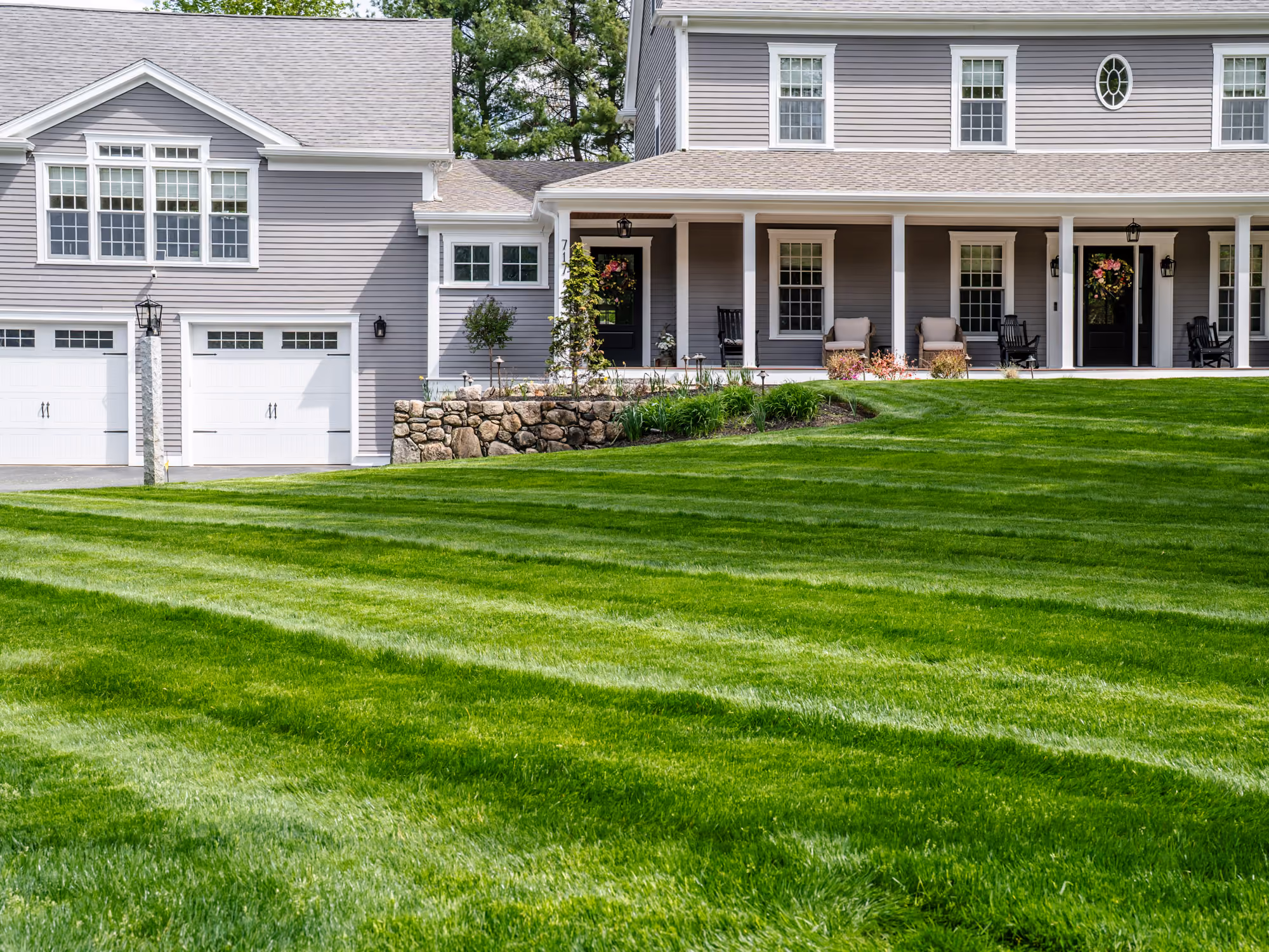 Backyard with a stone patio, small wooden deck with white railing, green lawn, and mulched garden areas with trees and shrubs.