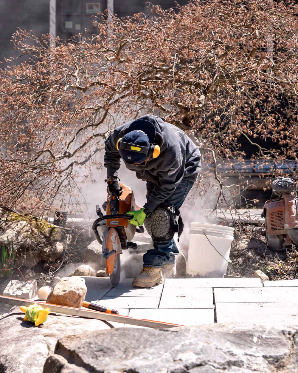 Partially completed stone patio and walkway under construction with stacks of paving stones, gravel, and a small orange skid steer loader nearby.