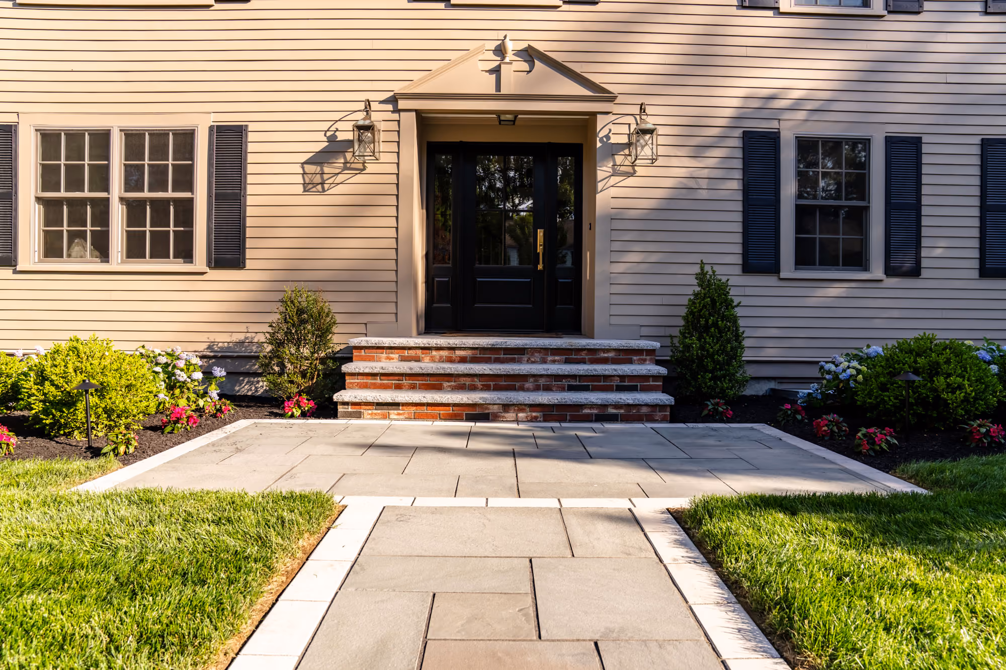A backyard with a newly paved stone patio, surrounded by garden soil, plants, and trees, adjacent to a house with a railing.
