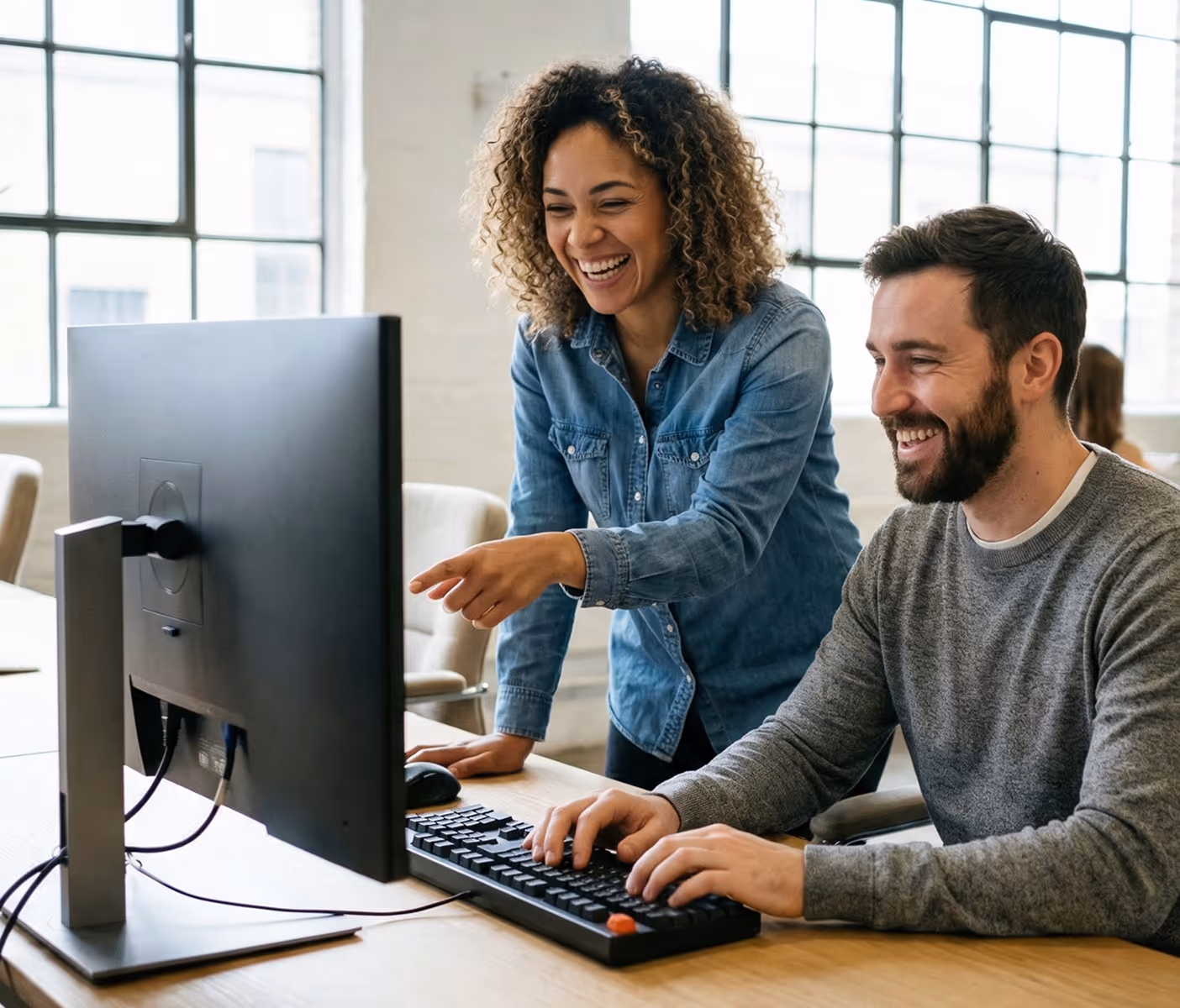 Smiling woman in denim shirt pointing at computer screen while man in gray sweater types on keyboard in office.